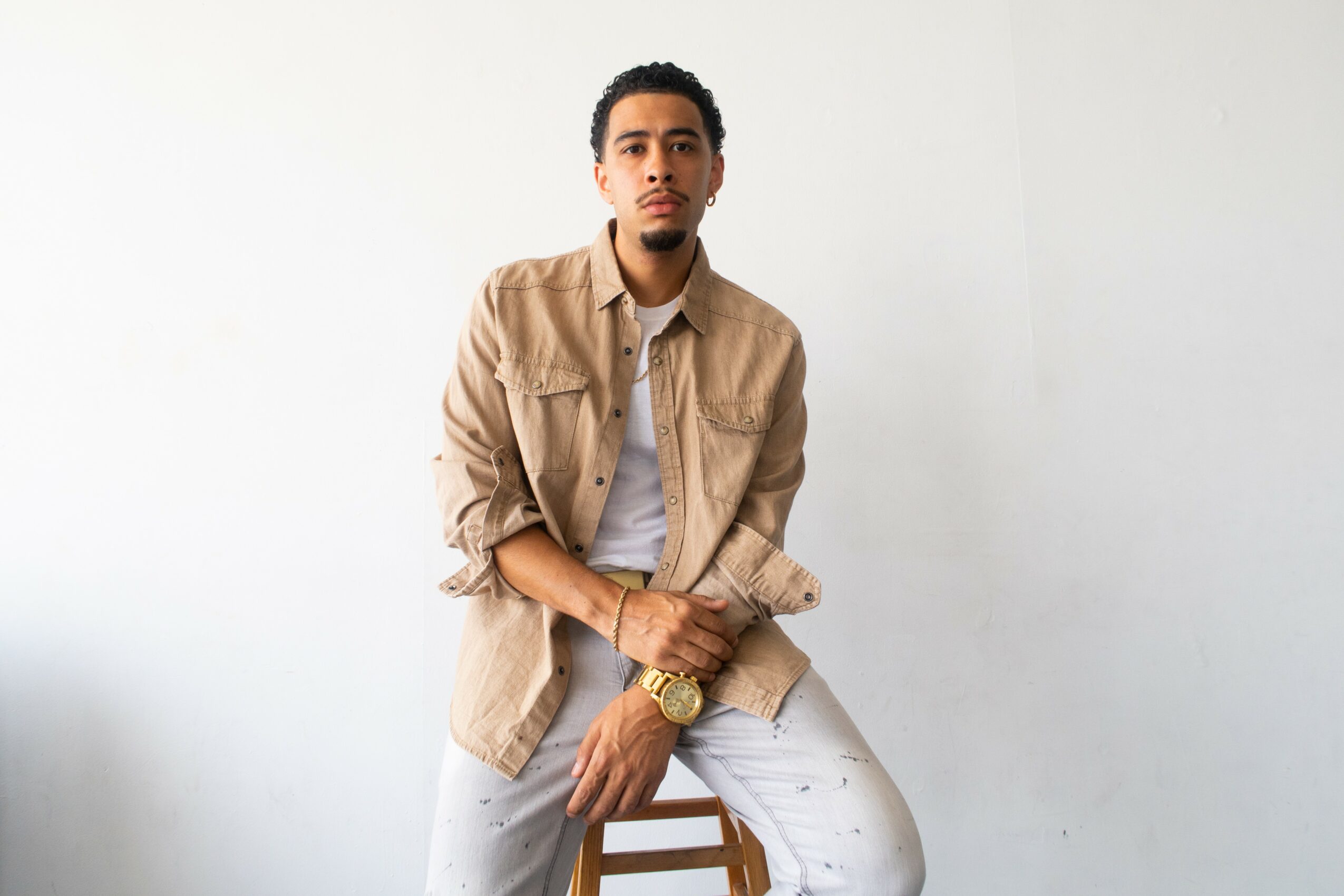 Young man sitting on a stool against a plain white wall, wearing a beige jacket and white pants.