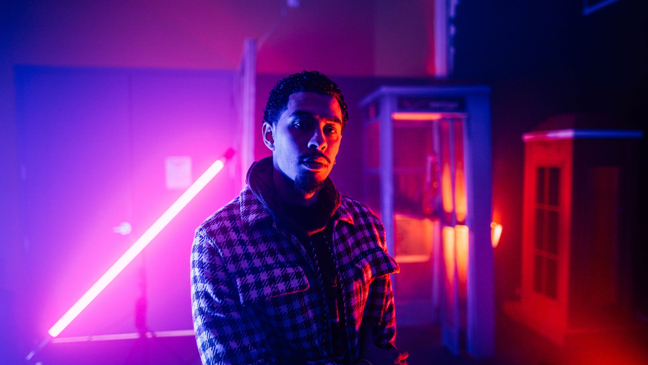 Person with short dark hair wearing a checkered shirt in colorful neon-lit environment.