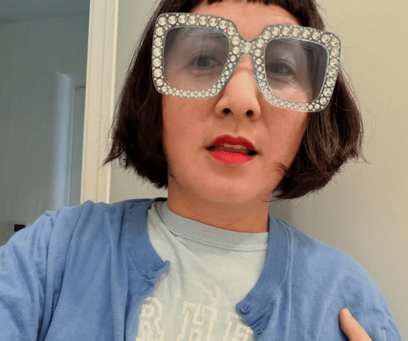 Person with short dark hair wearing large, rhinestone-encrusted glasses and a blue cardigan, looking at the camera.