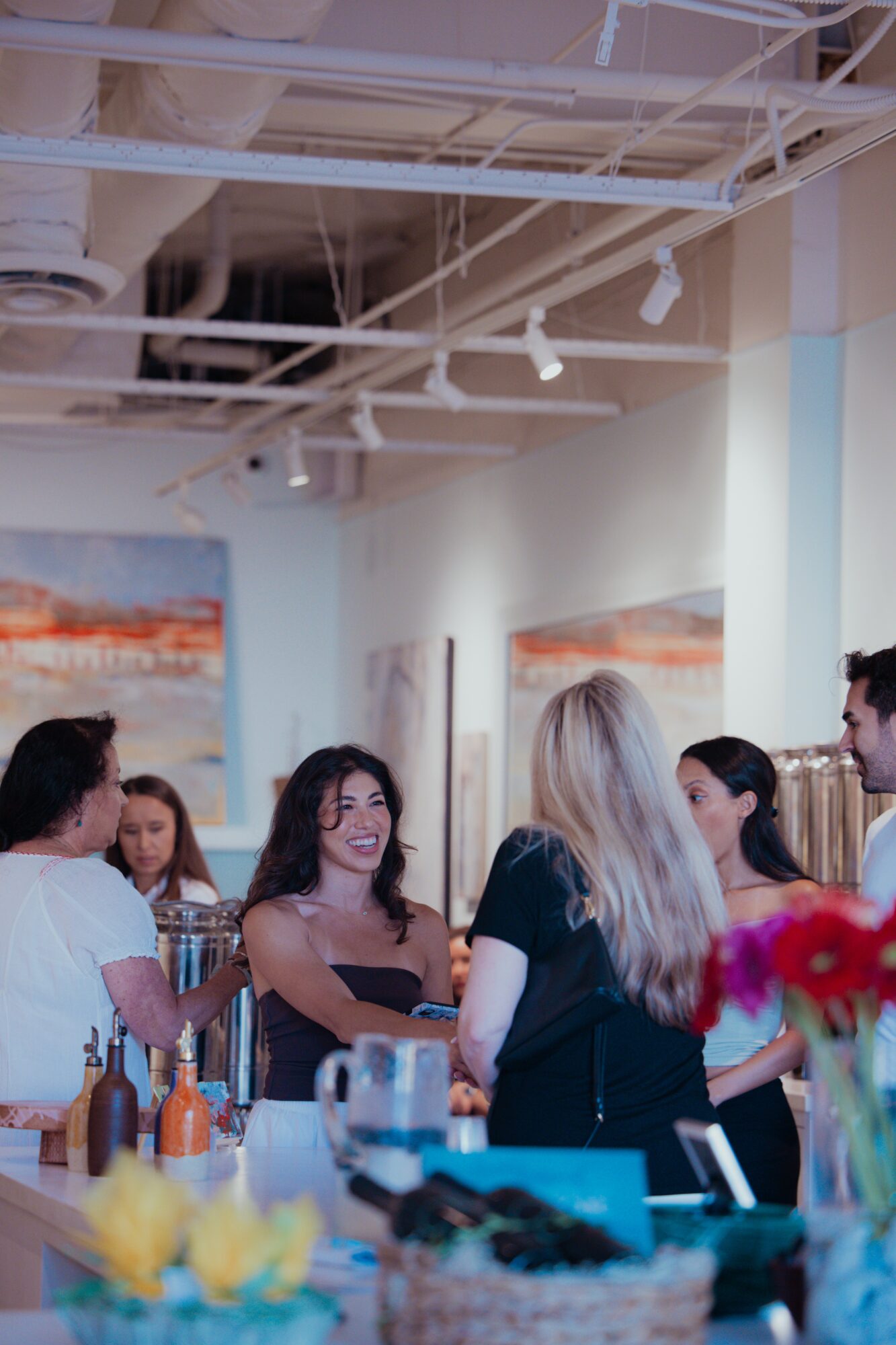 Group of people talking and smiling in an art gallery with paintings on the wall, colorful flowers in the foreground.
