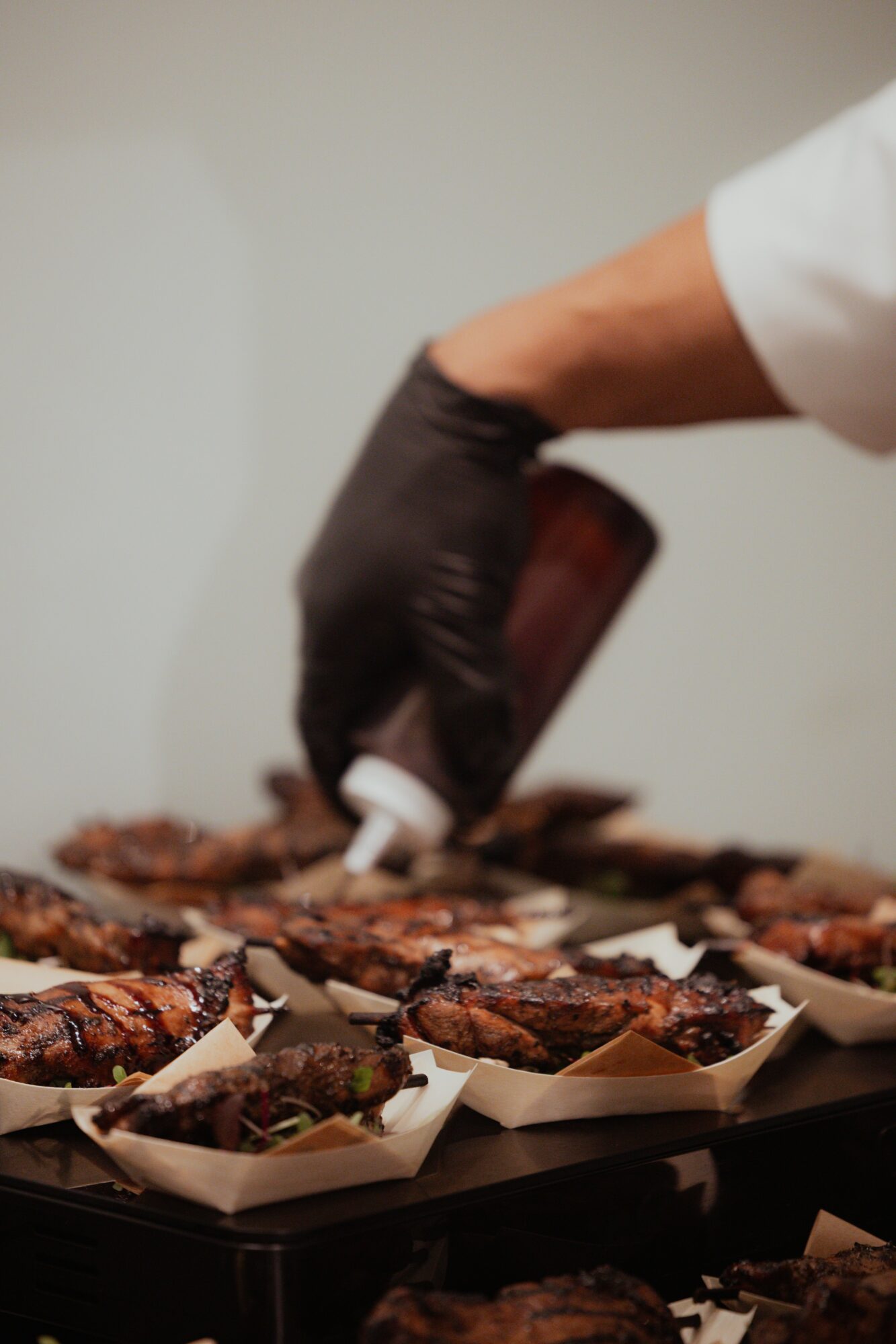 Person wearing black glove applying sauce to grilled meat on a tray, with multiple pieces of meat in paper trays.