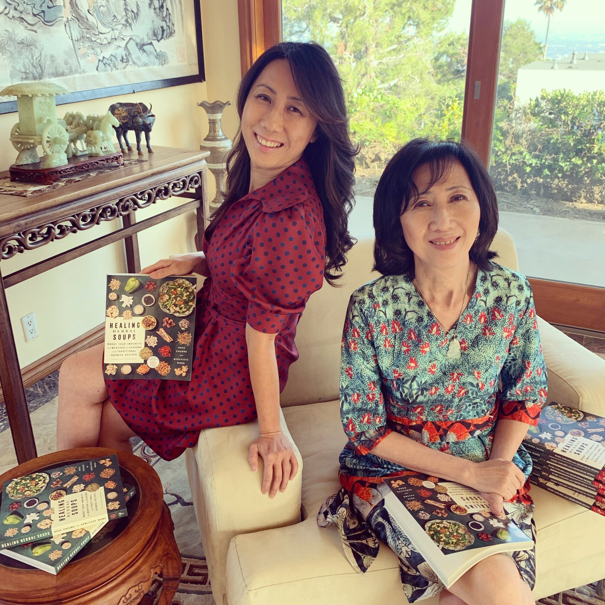 Two women sitting on a sofa, smiling, with books on their laps and a table nearby, in a room with large windows and greenery outside.