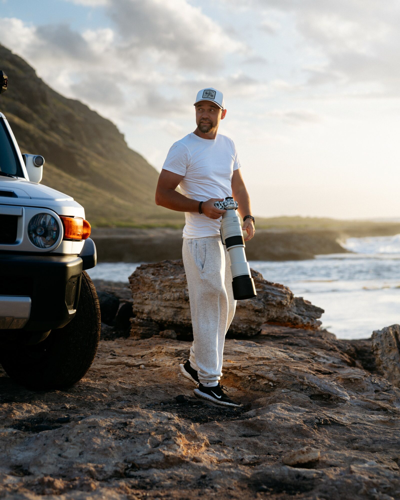 Man holding a camera with a large lens stands on rocky terrain near the ocean, with a vehicle nearby and a cloudy sky.