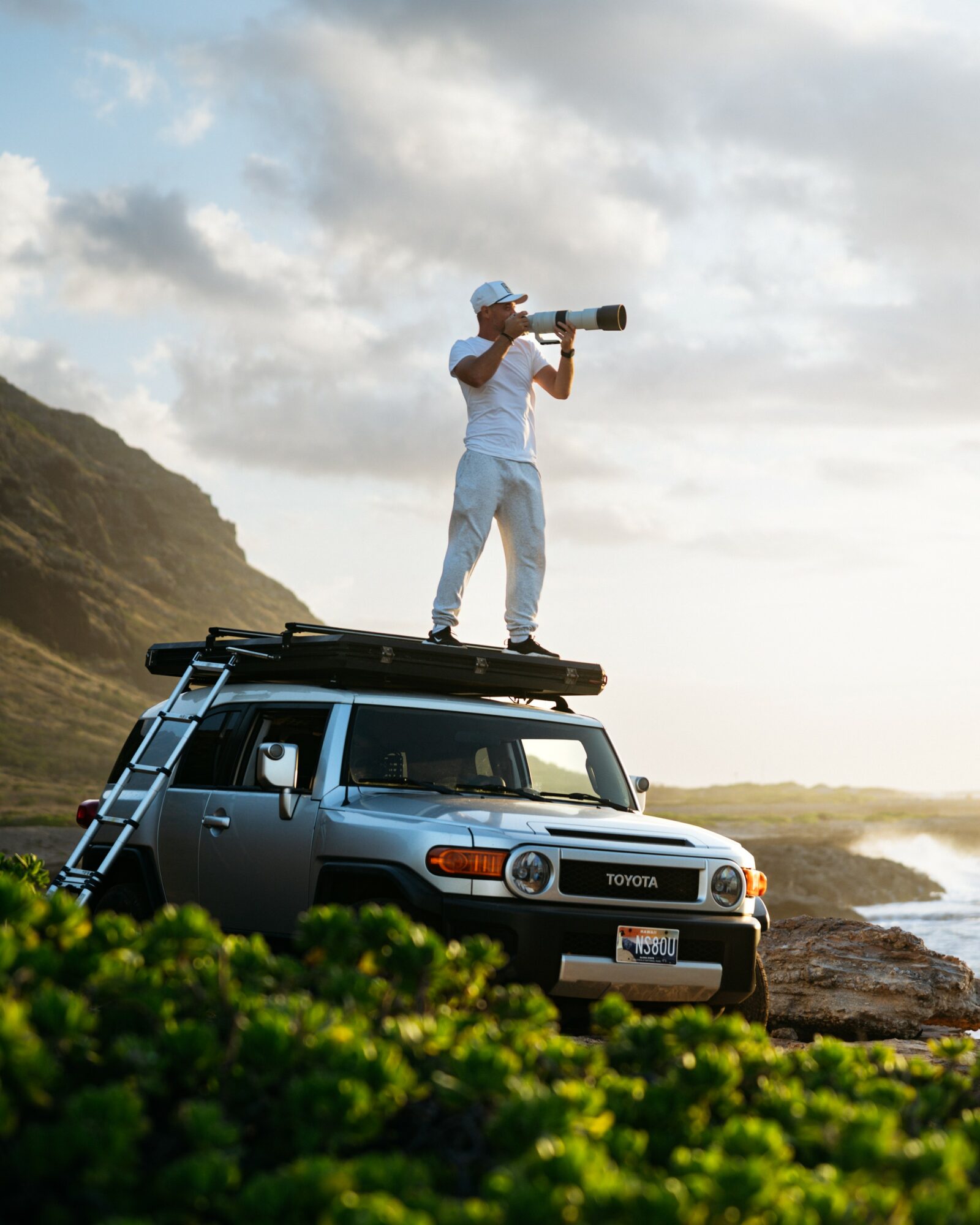 Person standing on a vehicle's roof, holding a telescope, near a rocky coastline with cloudy sky.