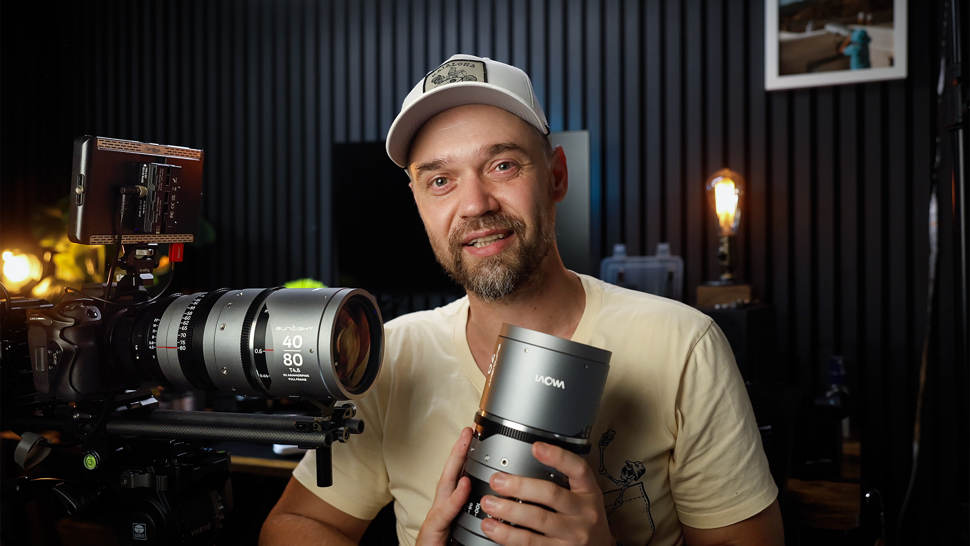 Man holding camera lens in a dark room with professional camera equipment, smiling at the camera.