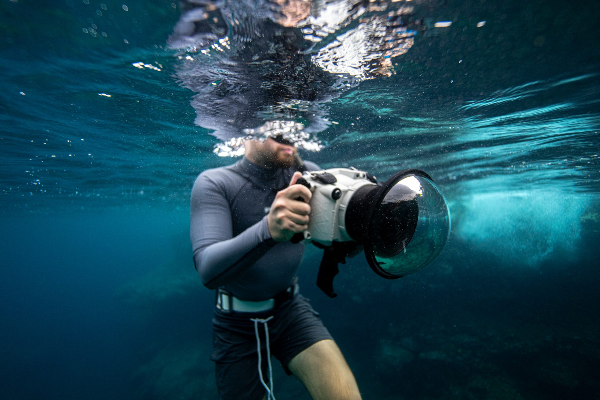 Person underwater holding a camera, wearing a wetsuit, with water and bubbles around them.