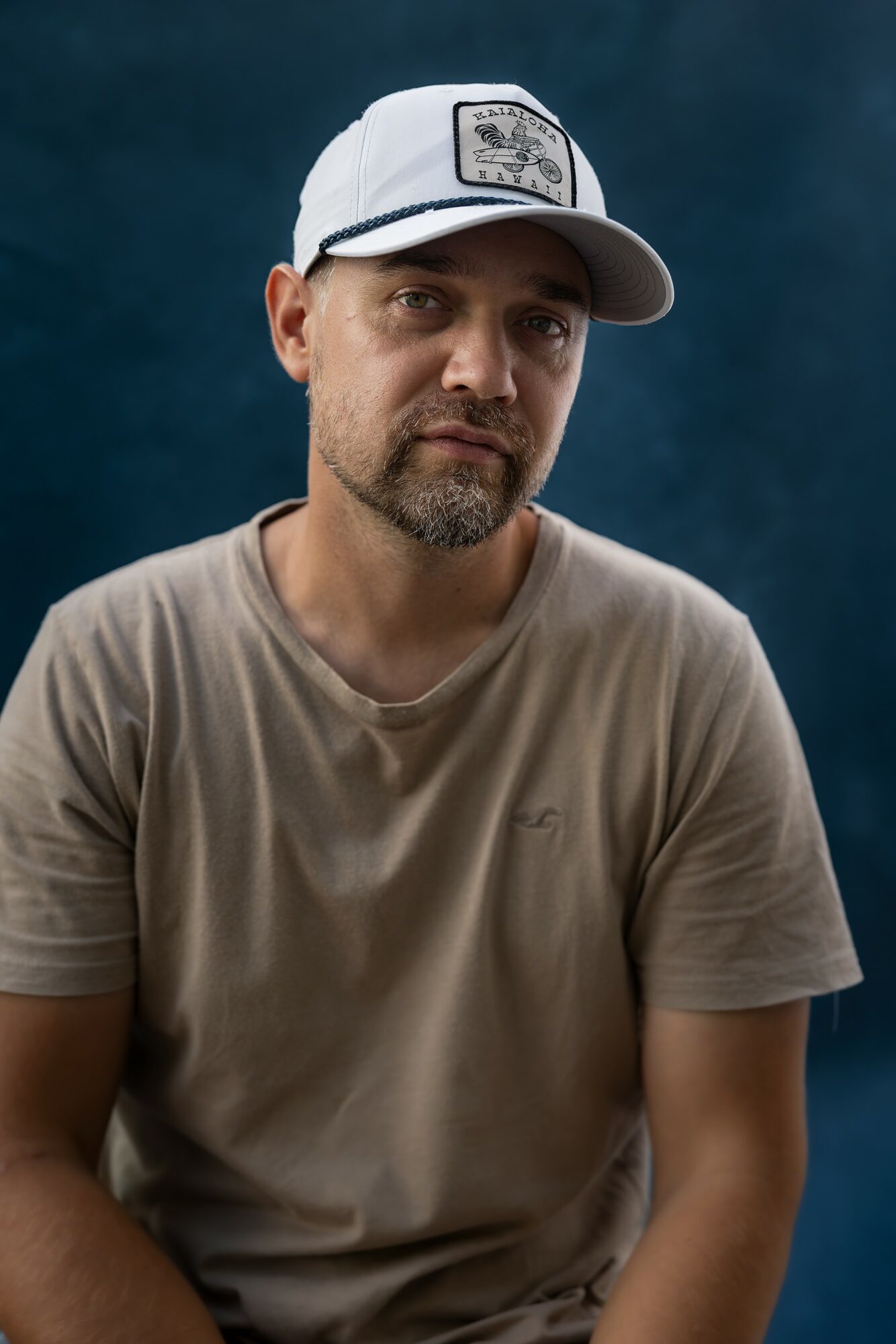 Man with a beard wearing a white cap and beige t-shirt, looking at the camera.