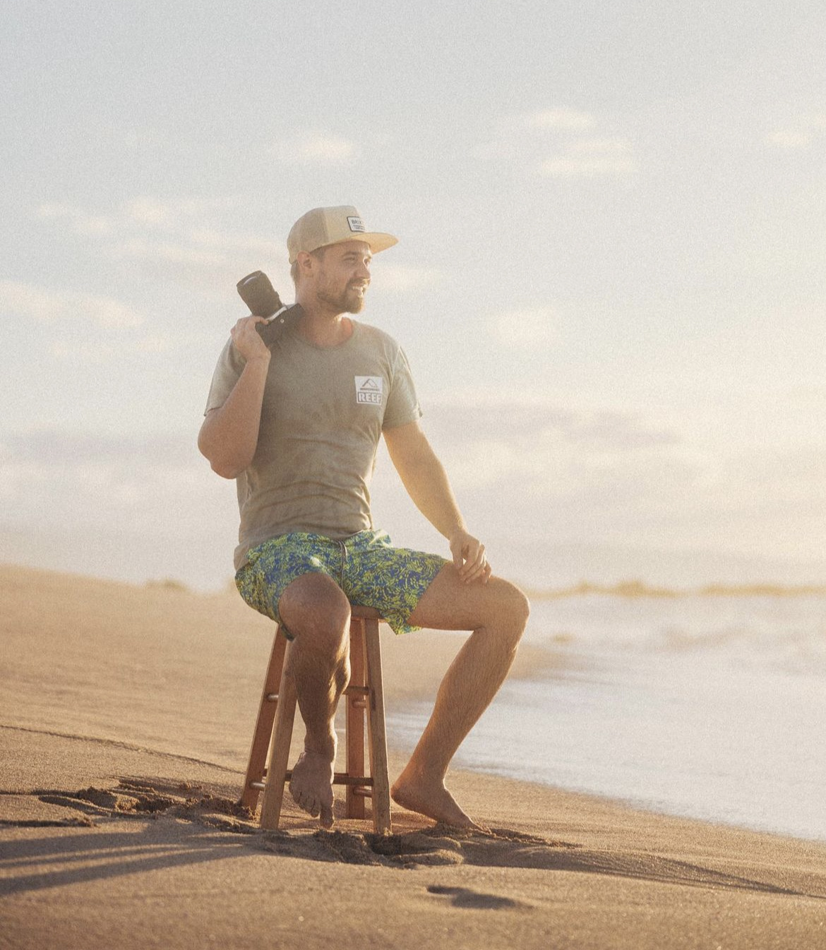 Man sitting on a stool on the beach, holding a camera, looking at the ocean, wearing a cap and casual clothes.