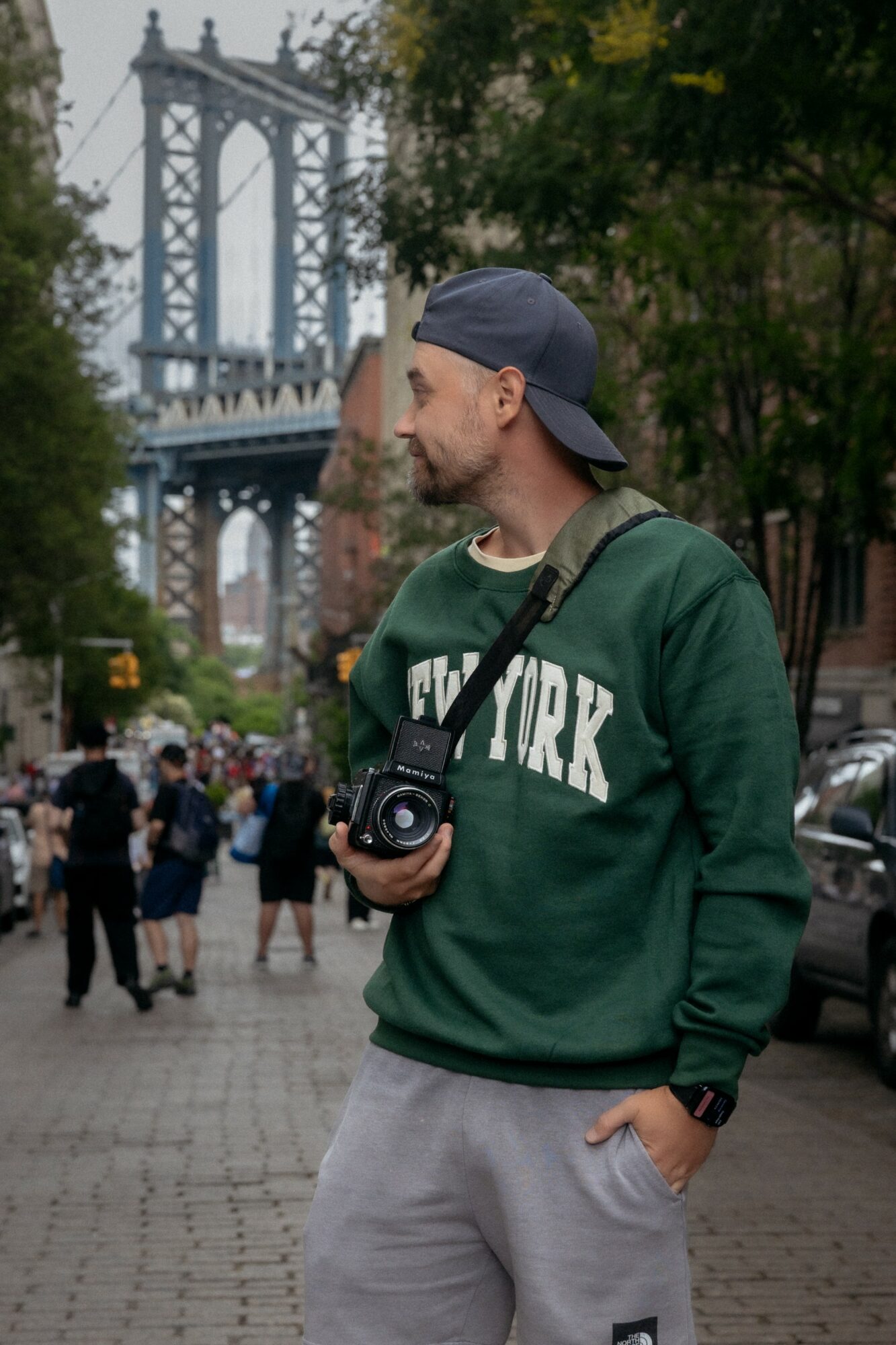Man in green sweatshirt and gray cap holding camera on city street with bridge in background.