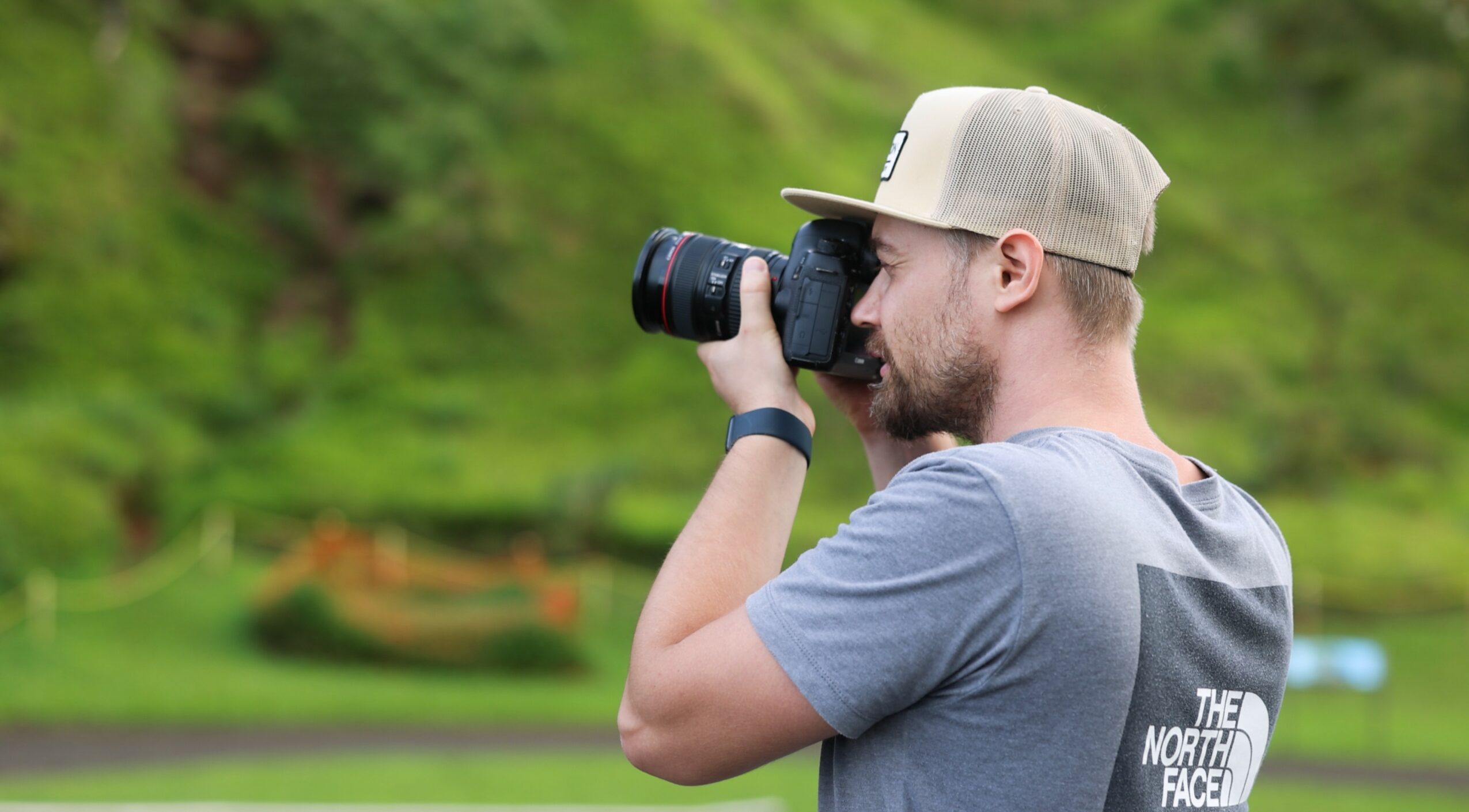 Man with a camera taking a photo outdoors, wearing a cap and gray T-shirt, with greenery in the background.