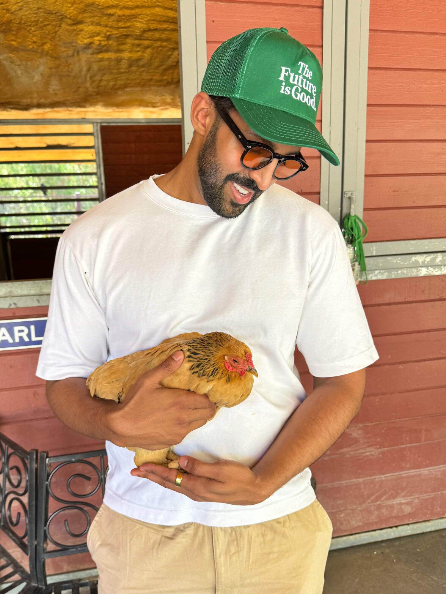 Man holding a chicken outdoors, wearing glasses, a green cap, and a white t-shirt, smiling.