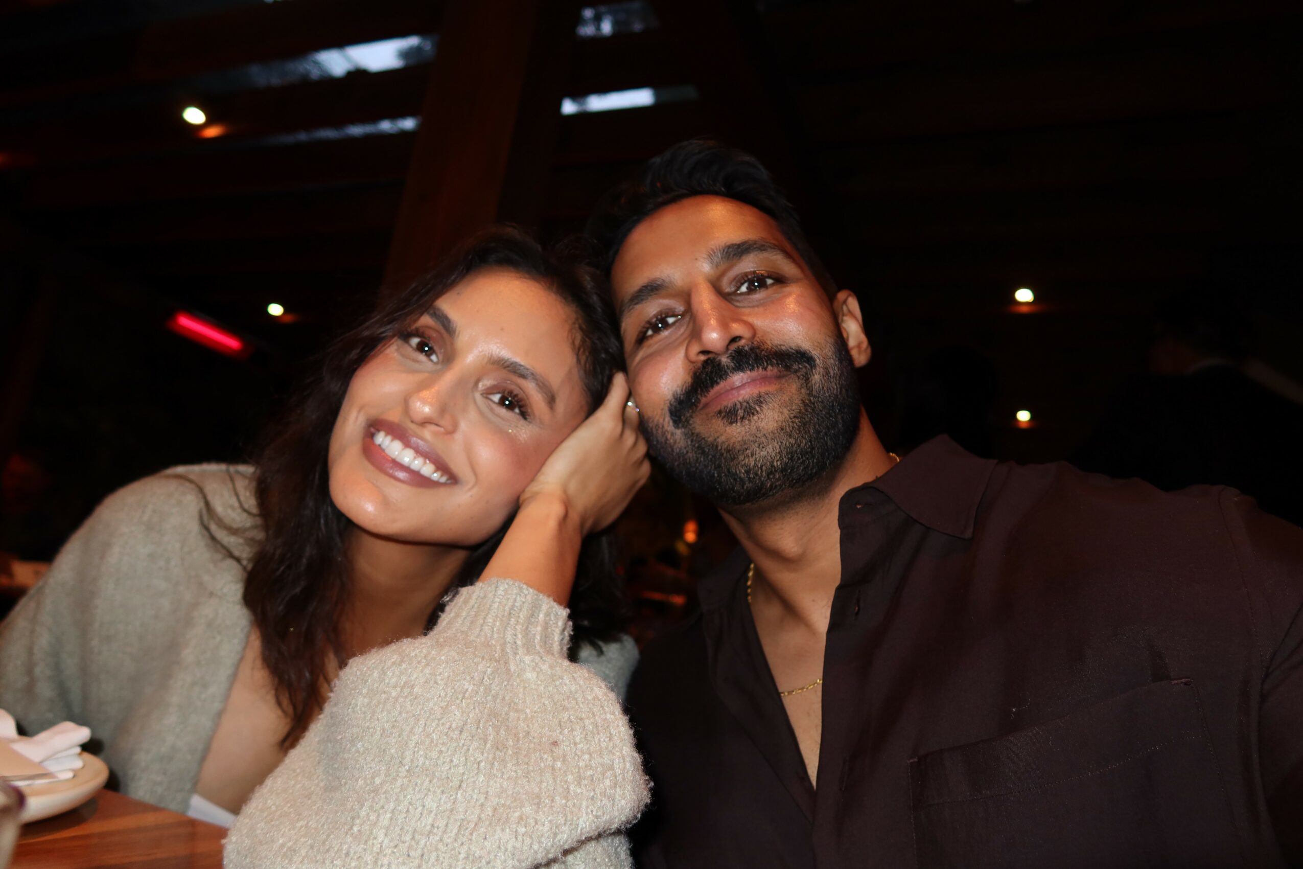 Two people smiling, close together, in a dark indoor setting, with warm lighting, one woman and one man, both looking at the camera.