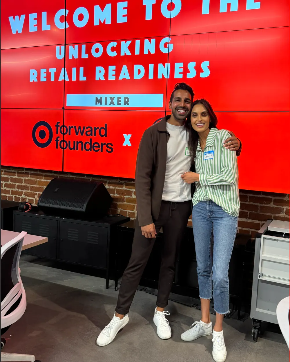 Two people smiling and standing together in front of a red digital display with text about retail readiness, in an indoor setting.