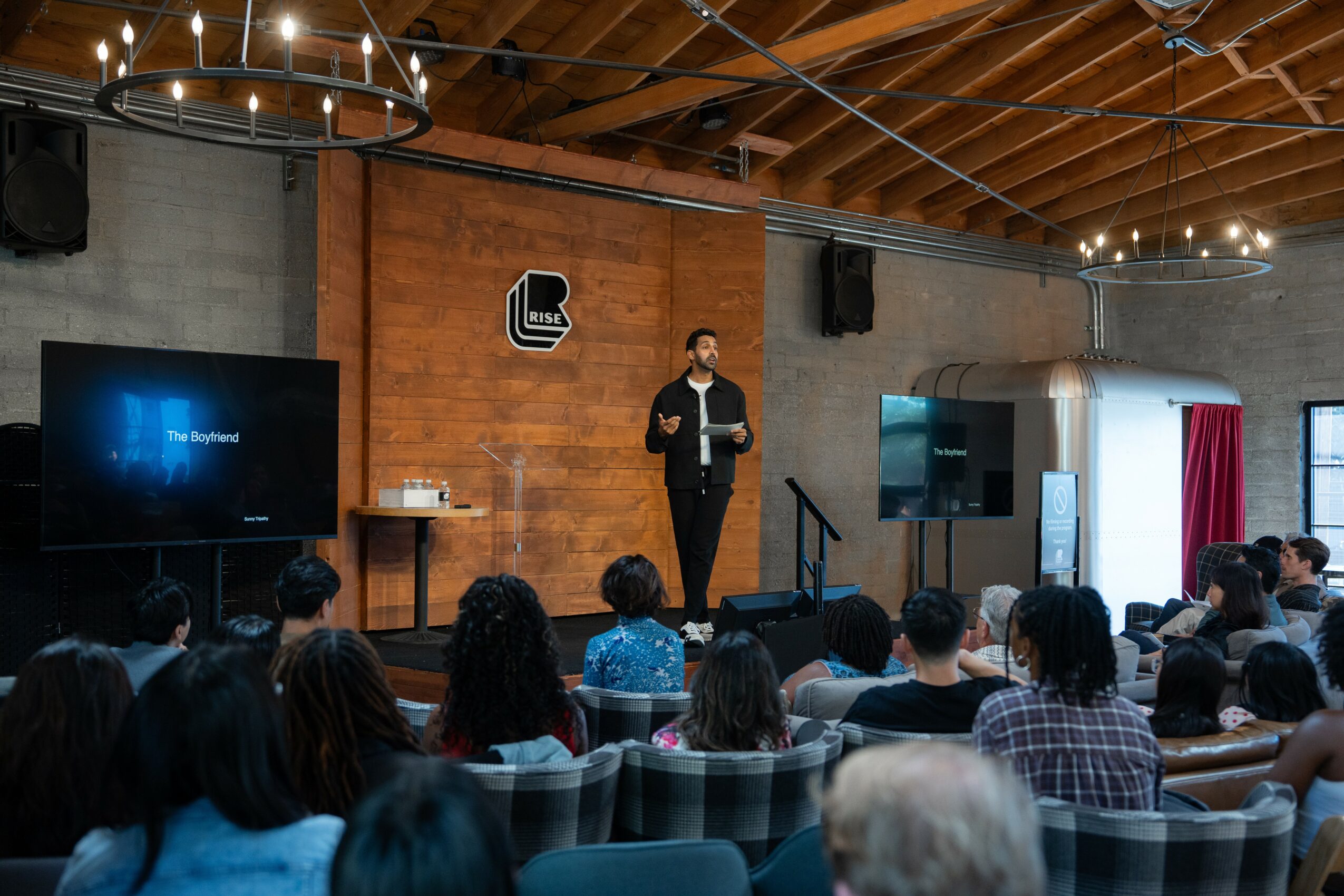 Person standing on stage giving a presentation to an audience in a room with brick walls and wooden ceiling, two large screens, and a chandelier.