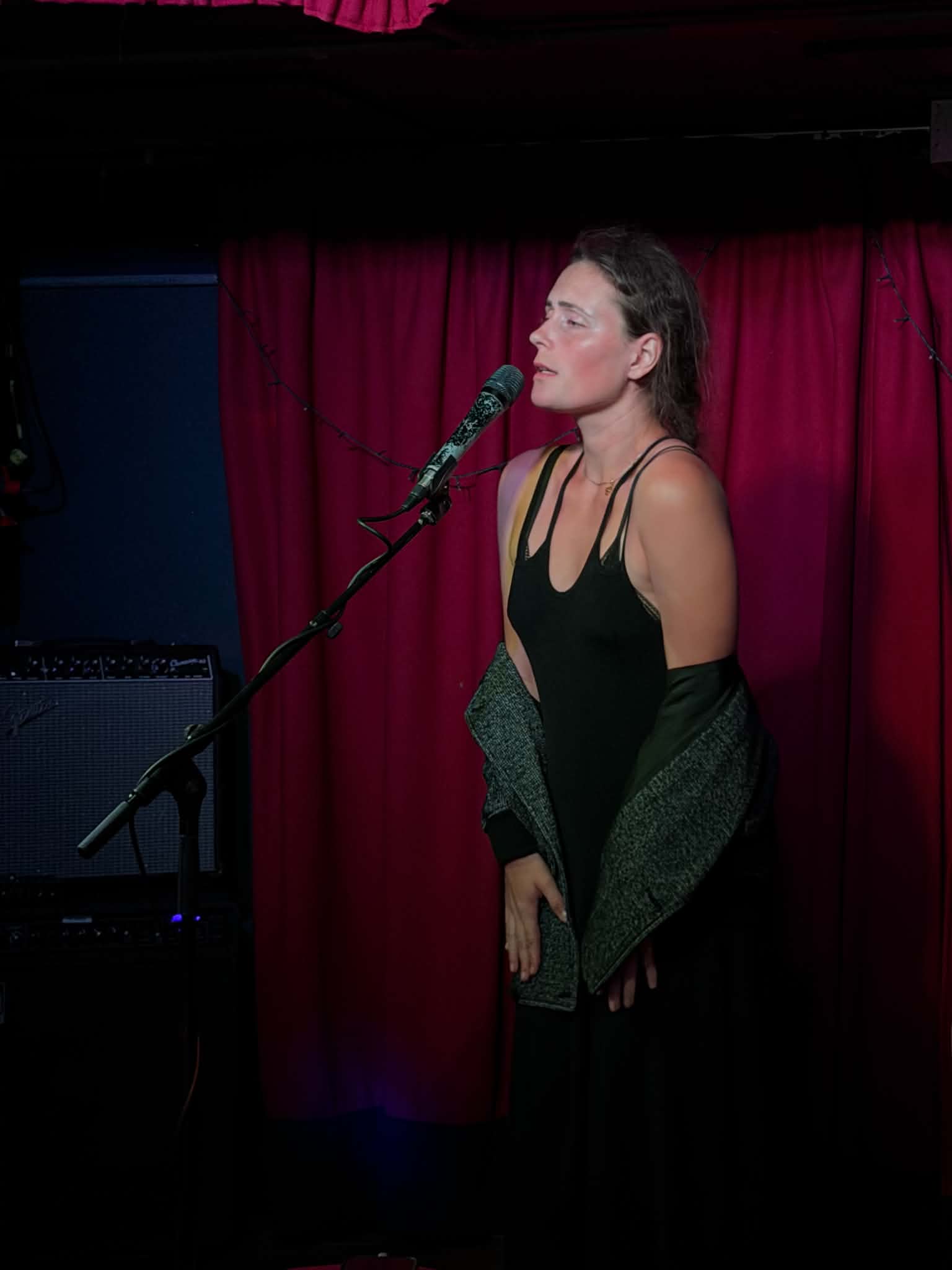Woman singing into a microphone on stage with red curtains background.