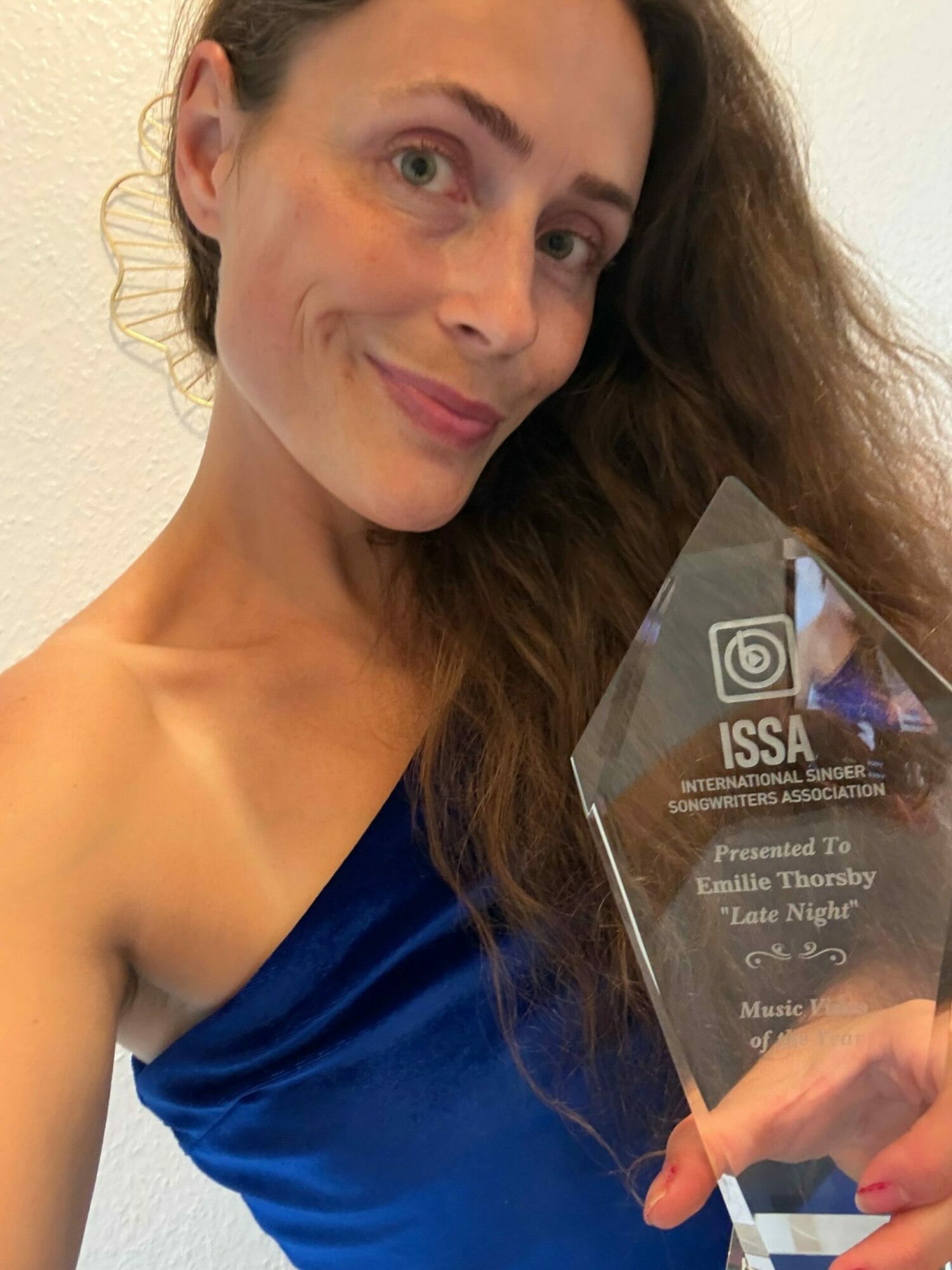 Woman holding a glass award, smiling, with long brown hair, wearing a blue top, against a plain background.