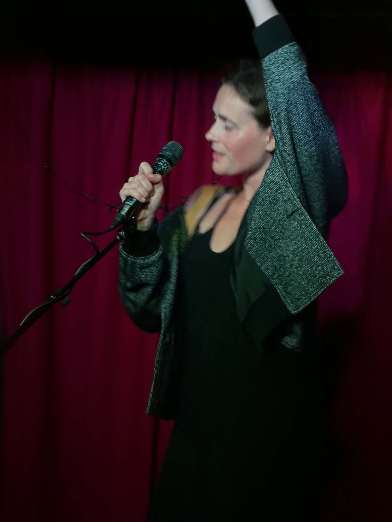 Woman holding a microphone with one arm raised, standing in front of a red curtain, wearing a dark dress and a green jacket.