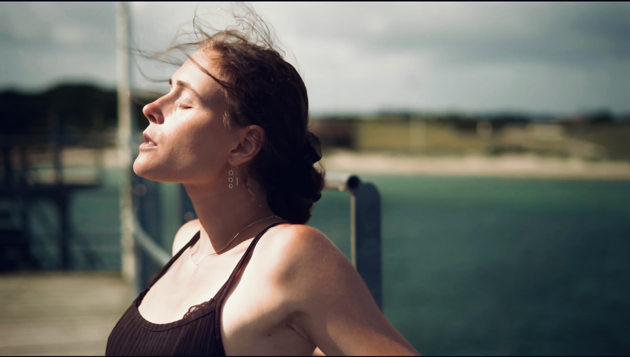 Woman with closed eyes outdoors near water, sunlight on face, dark top, hair pulled back.