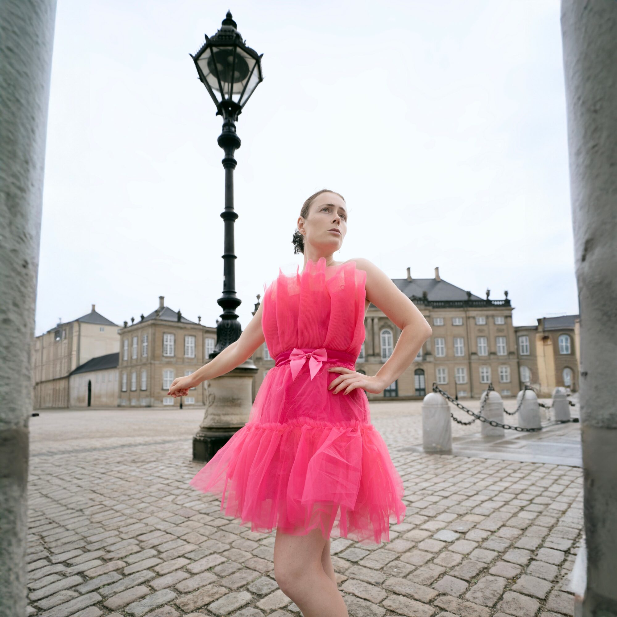 Woman in pink dress standing outdoors near a lamppost and historic buildings.