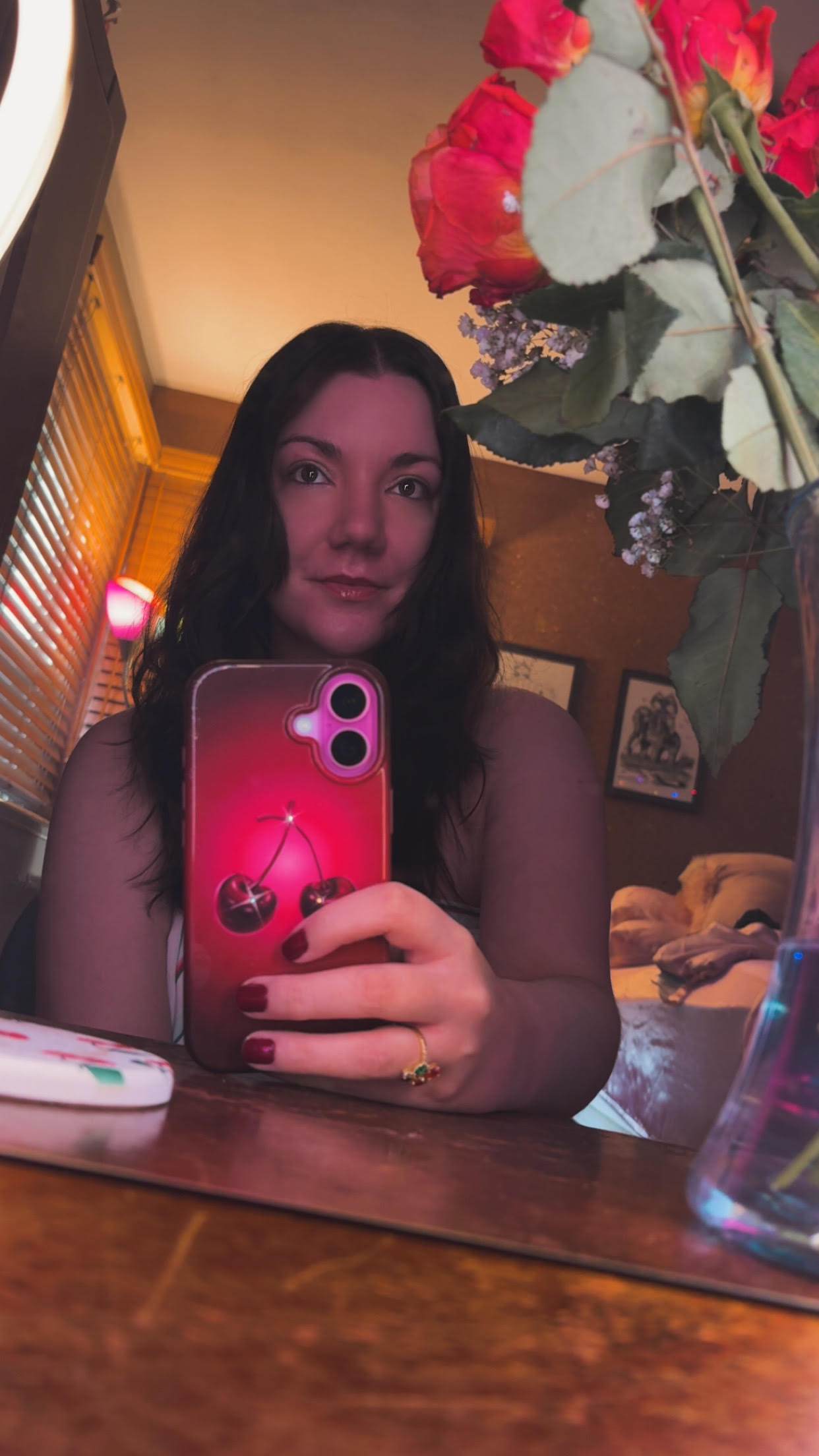 Woman with long dark hair taking a mirror selfie in a room with warm lighting, holding a pink phone, with a bouquet of flowers above.