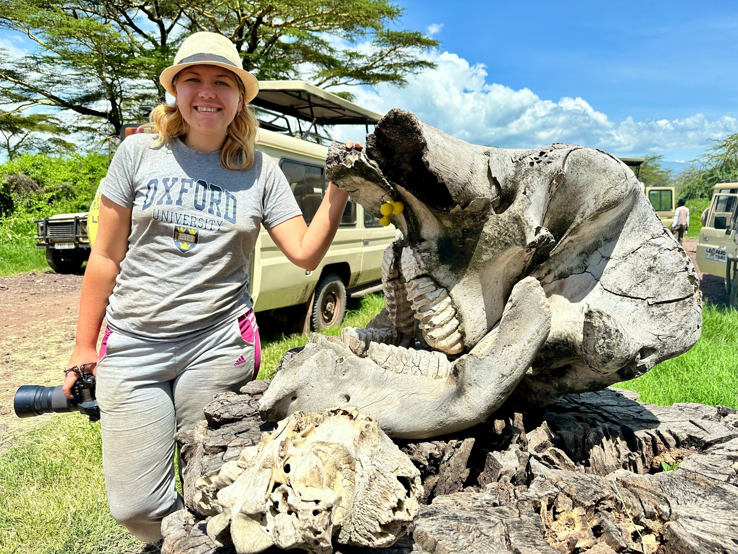 Woman smiling with a hat, holding a crocodile sculpture's mouth open outdoors, with trees and vehicles in the background.