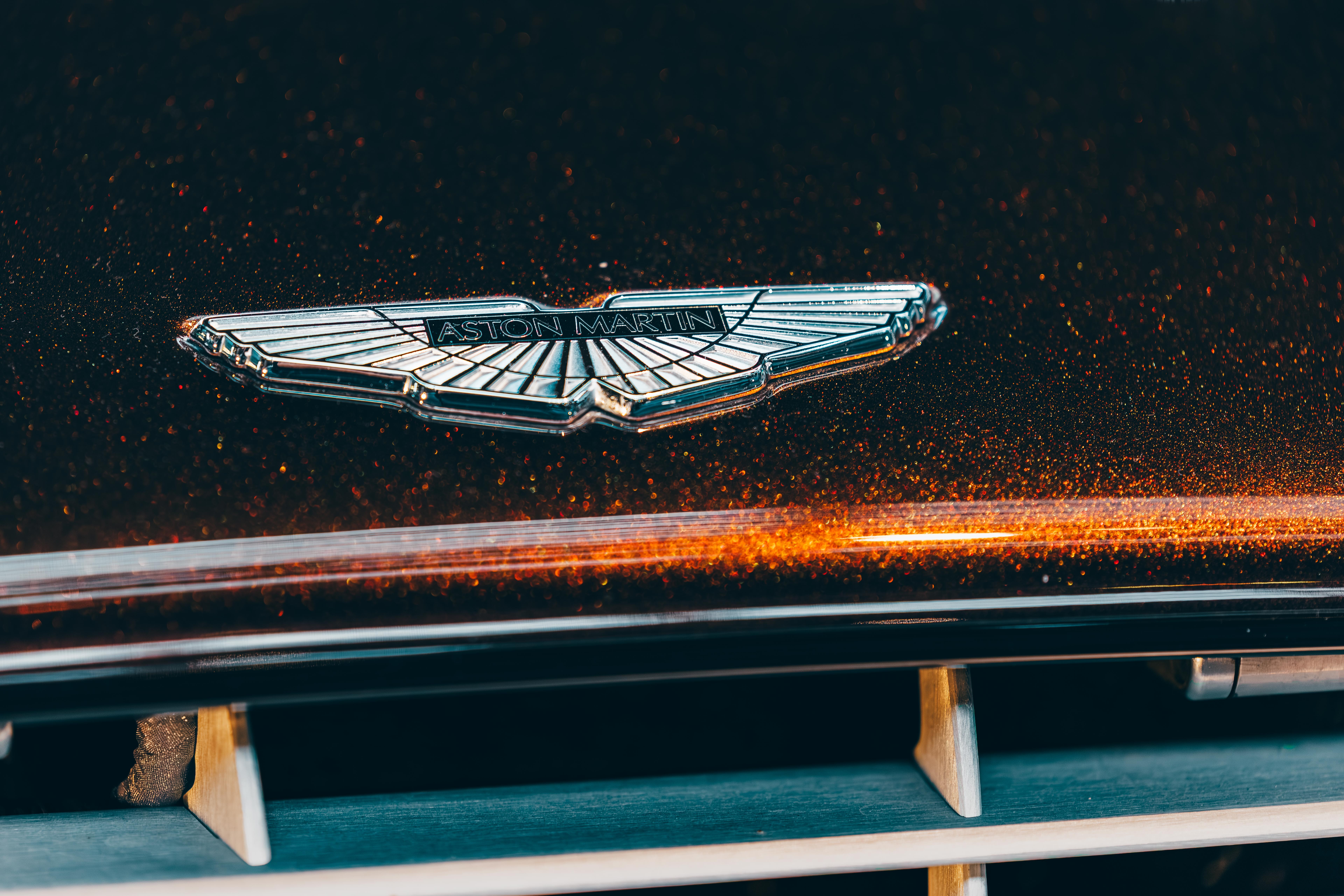 Close-up of an Aston Martin logo on a car's hood, with a dark background and orange light streaks.