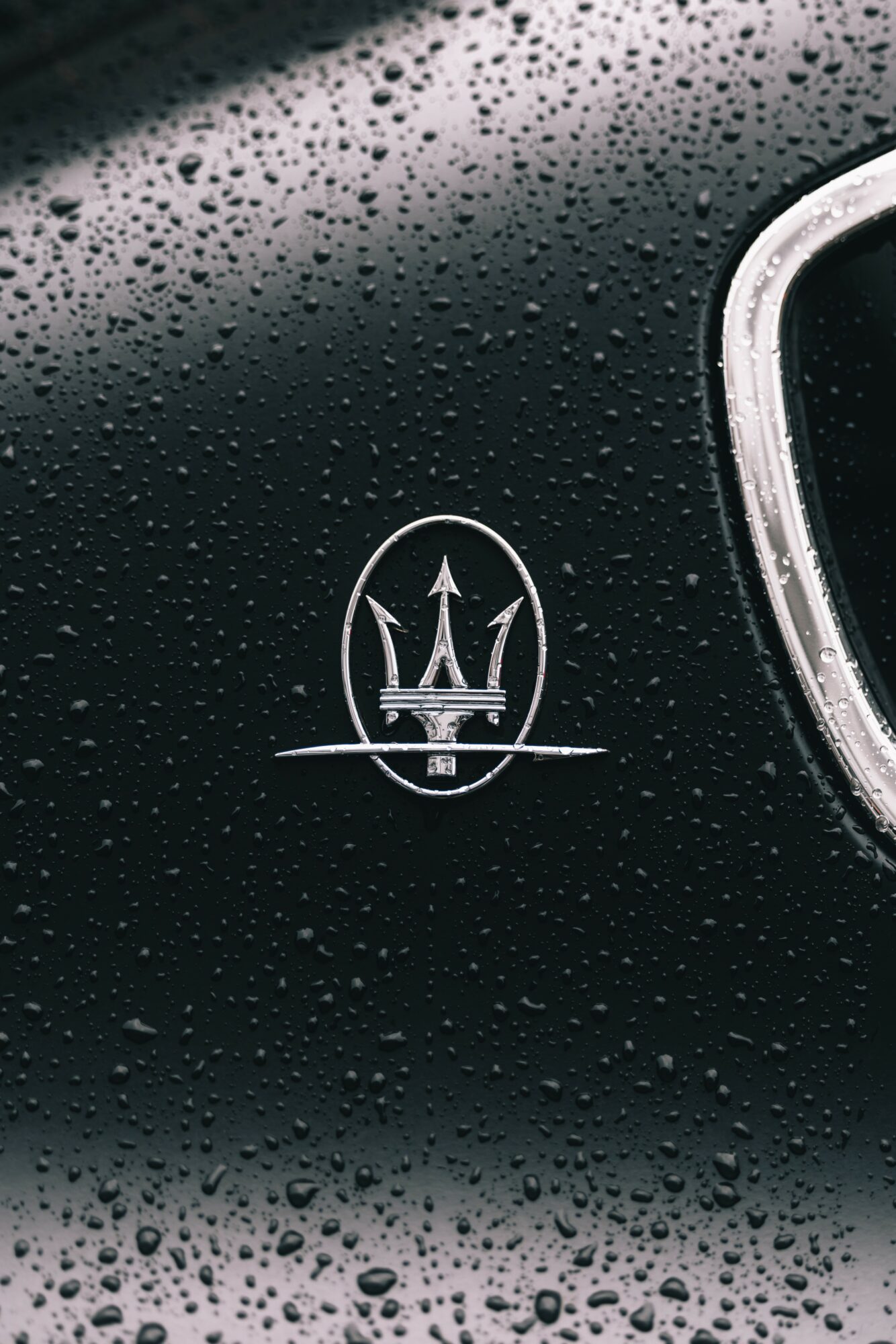 Close-up of a black surface with water droplets and a white Maserati logo, partially visible on the right.