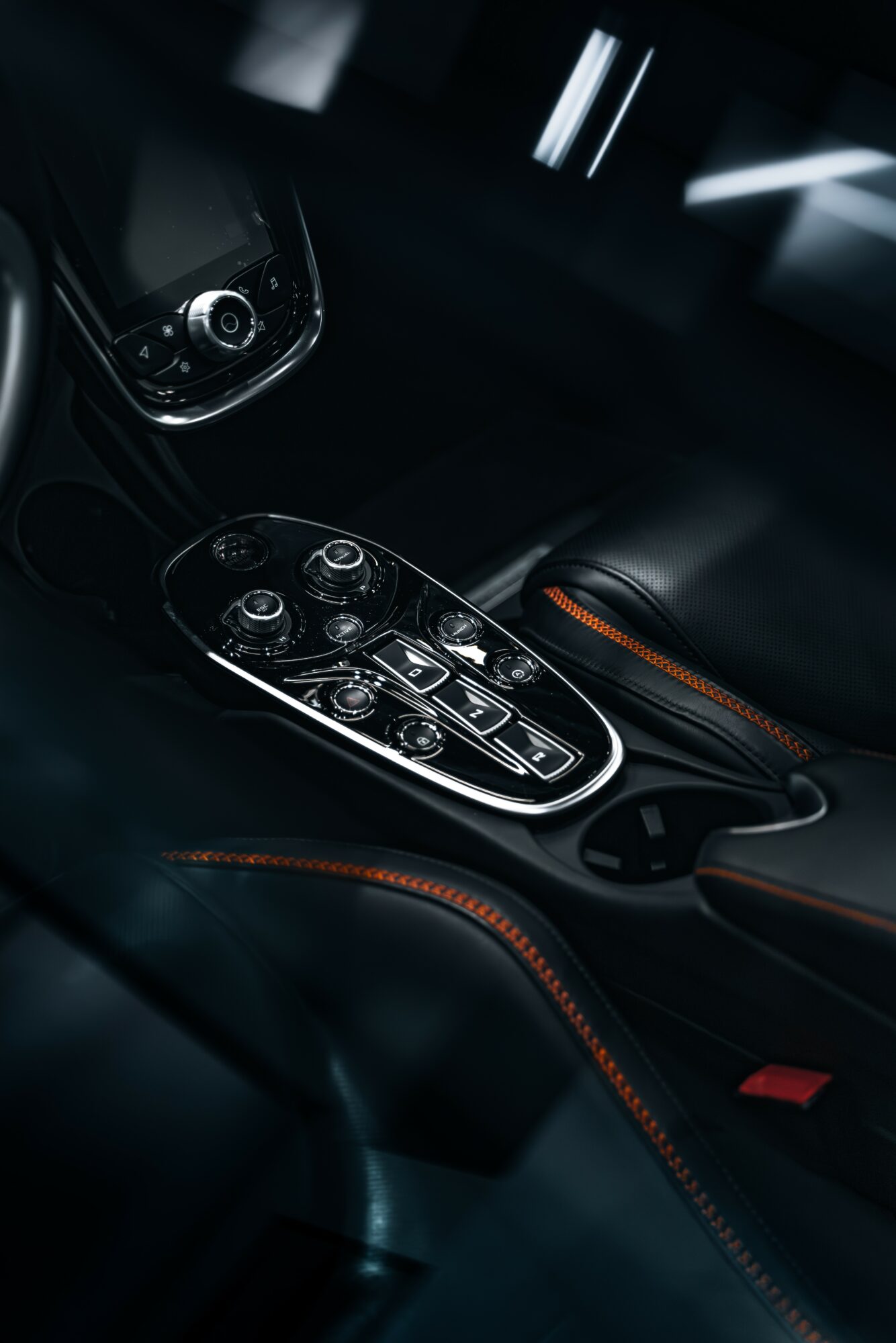 Close-up of car interior center console with control buttons and gear shift area, black with orange stitching, dim lighting.
