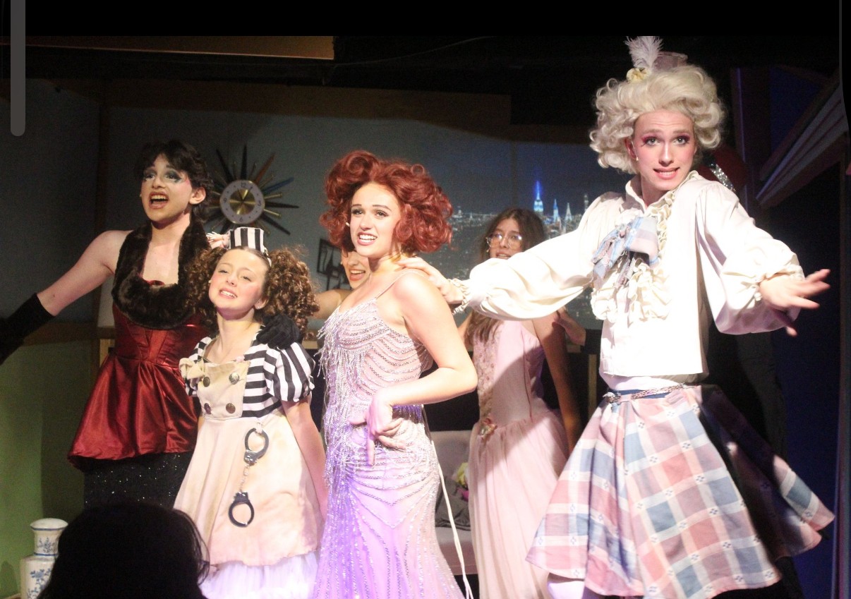 Group of women and children in colorful costumes on stage, smiling and posing, with a decorative background.