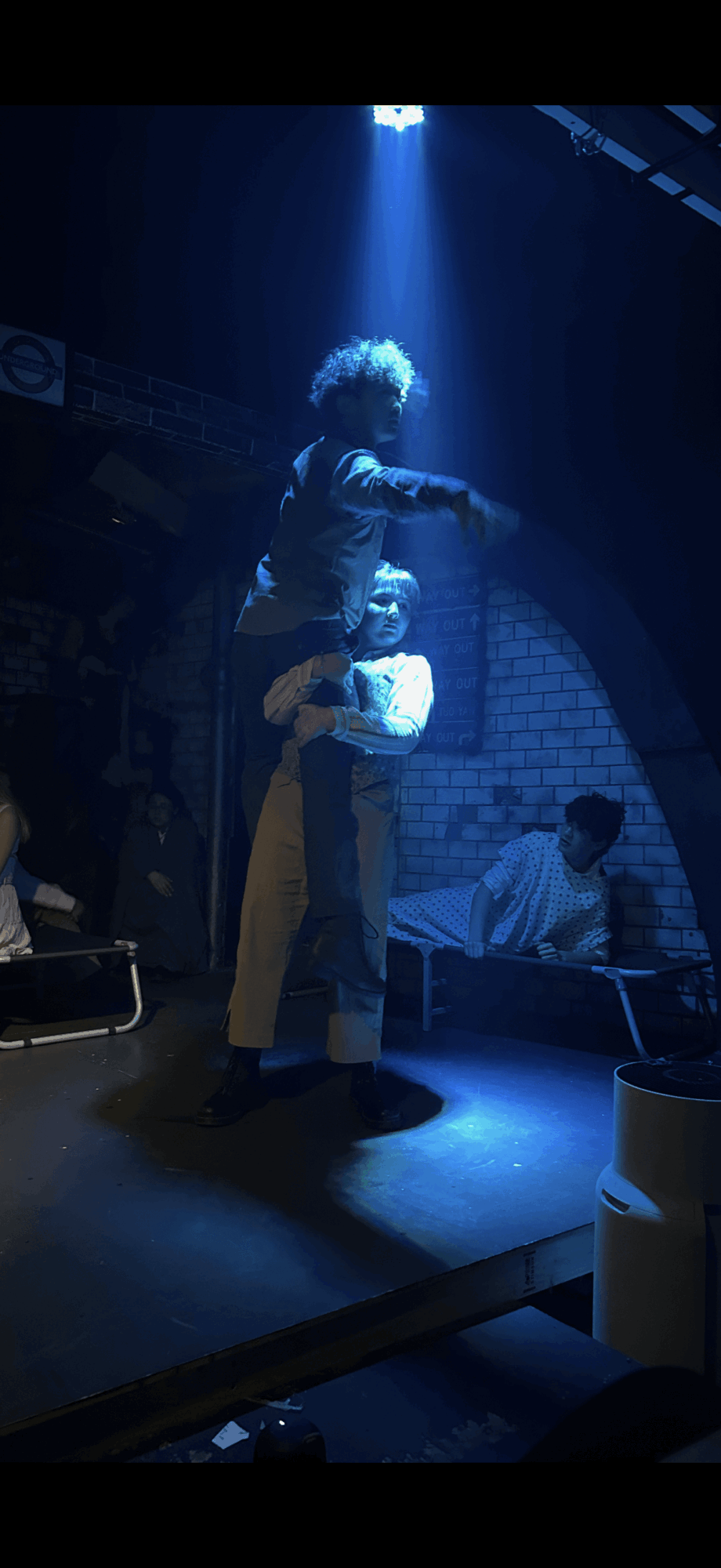 Performer with curly hair stands on stage under blue spotlight, with two seated people in the background.