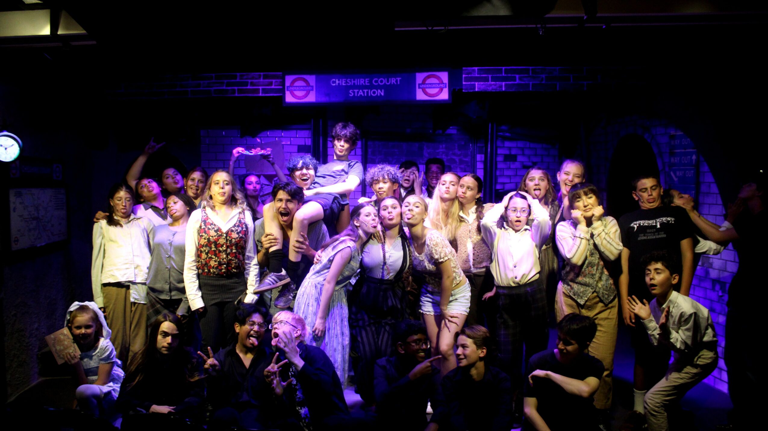 Group of people on stage, some posing, with a dark background and purple lighting, in a theatrical setting.