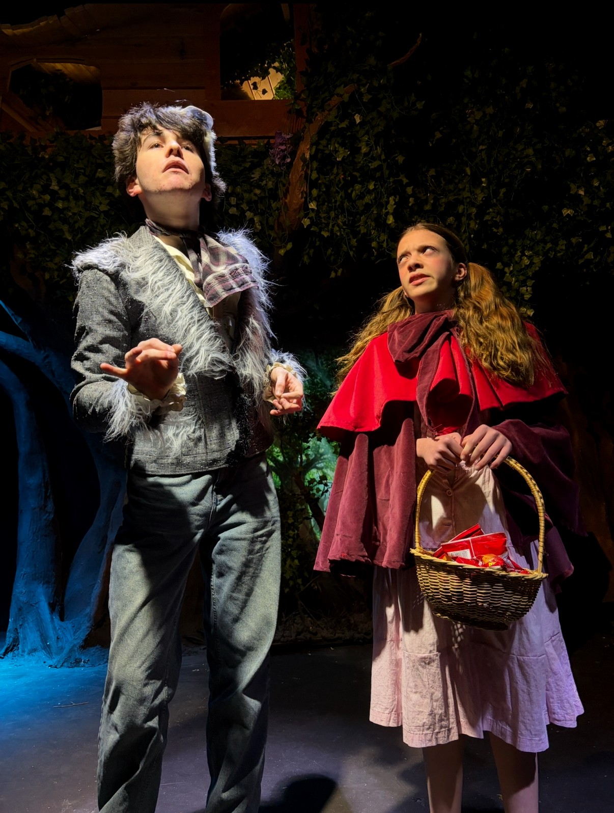 Two young women on stage, one with gray hair and a fur vest, the other with long hair and a red cape, holding a basket.
