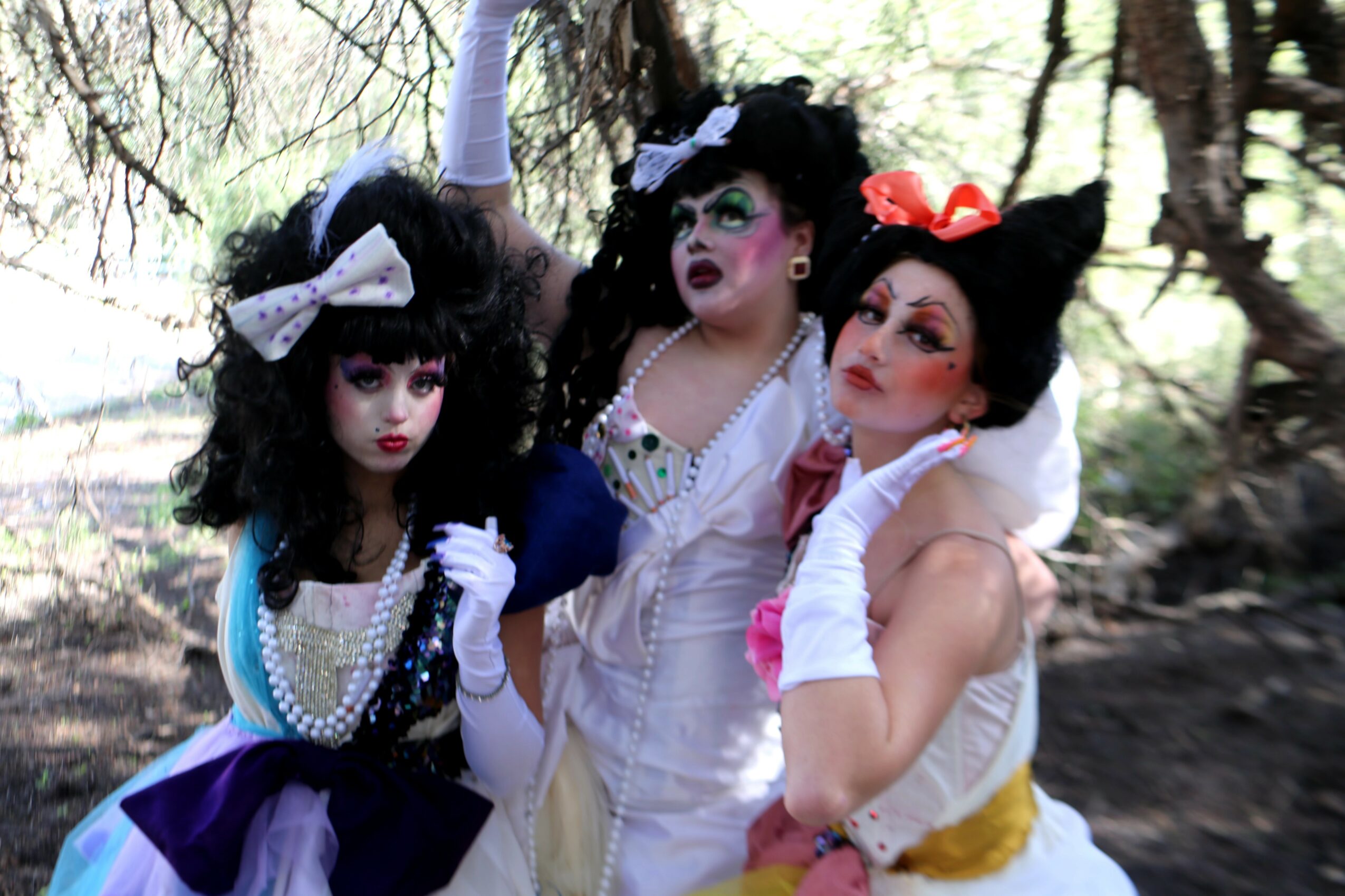 Three people dressed in elaborate costumes and makeup pose outdoors among trees and rocks.