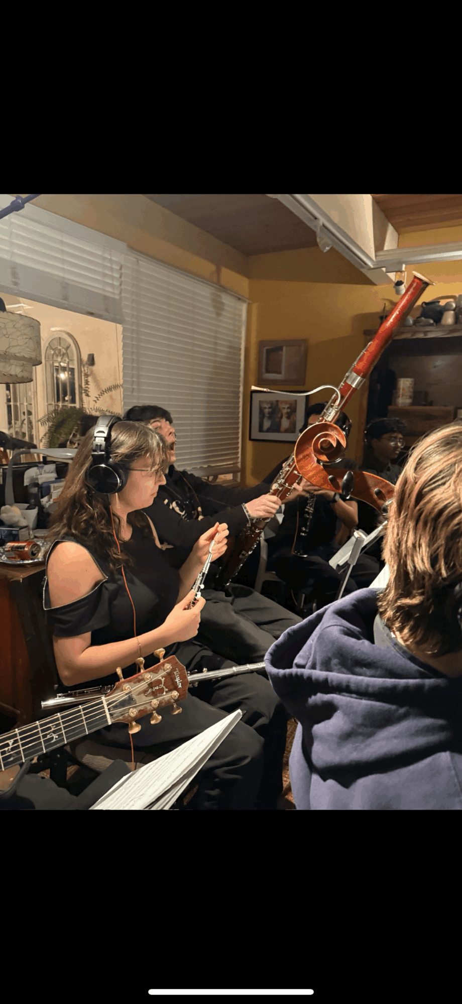 Group of people playing musical instruments indoors, including a person with a saxophone and another with a guitar, in a cozy room.