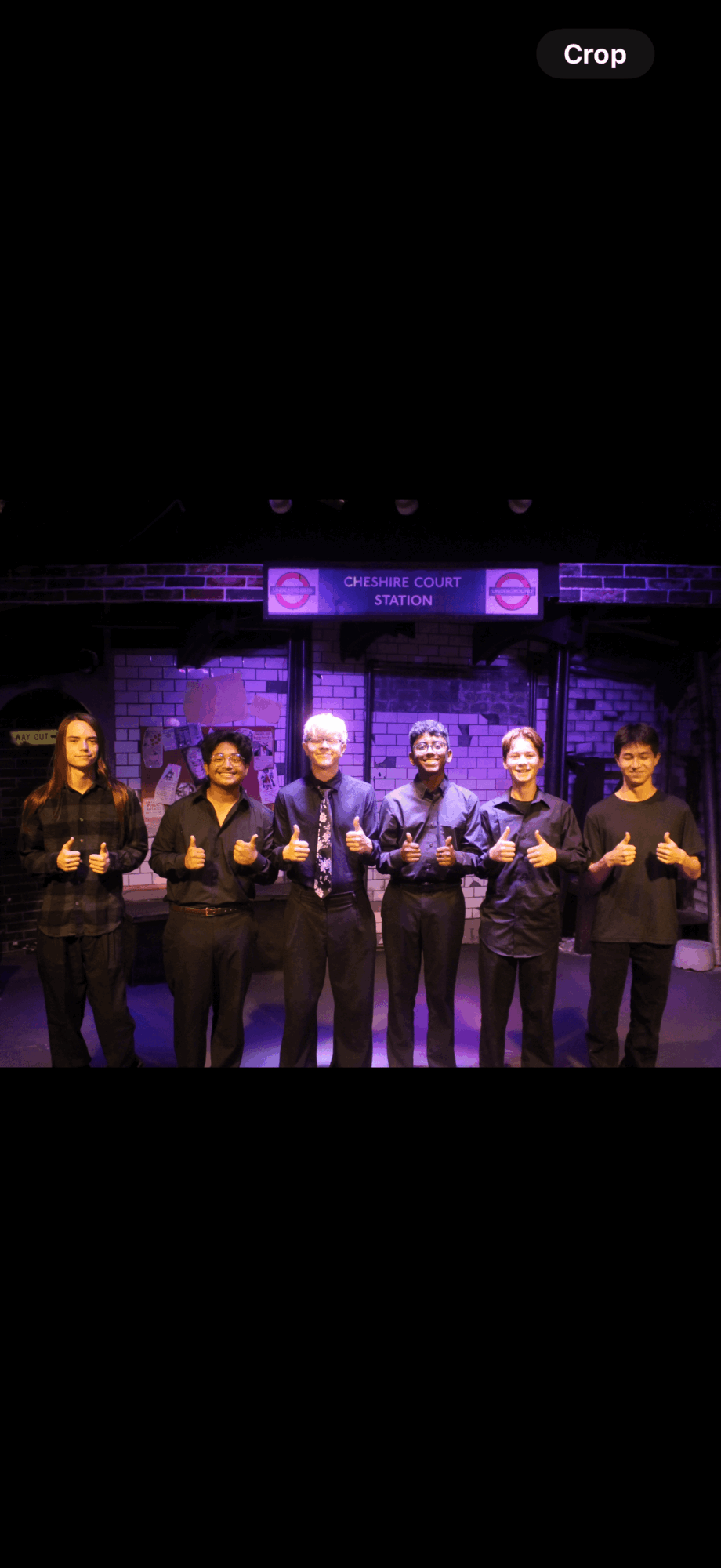 Six people stand on stage with a dark background and purple lighting, holding microphones and smiling.