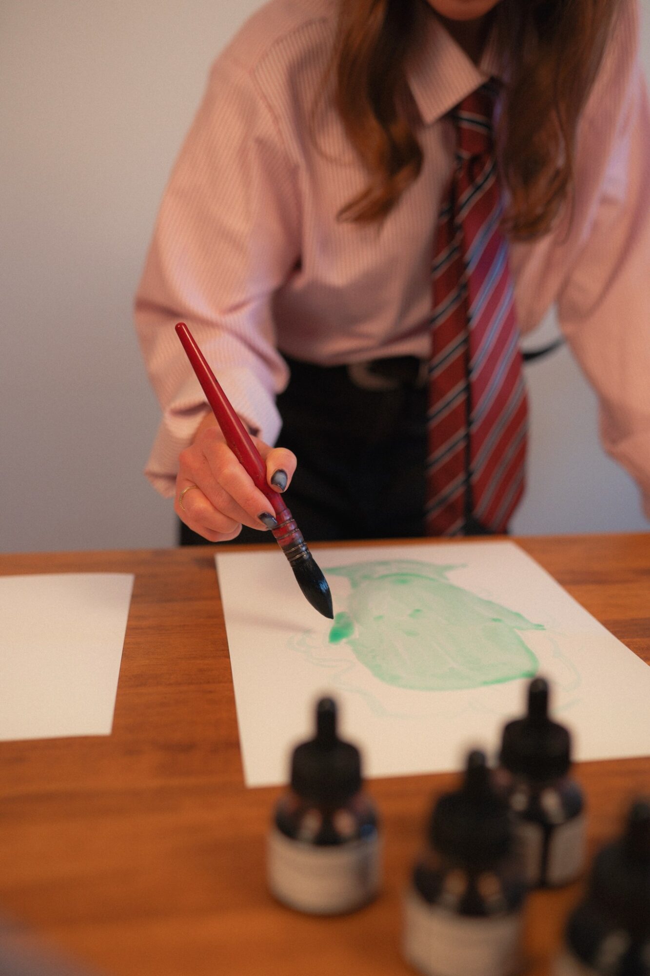 Person in school uniform painting on paper with watercolors, bottles of ink on table, face not visible.