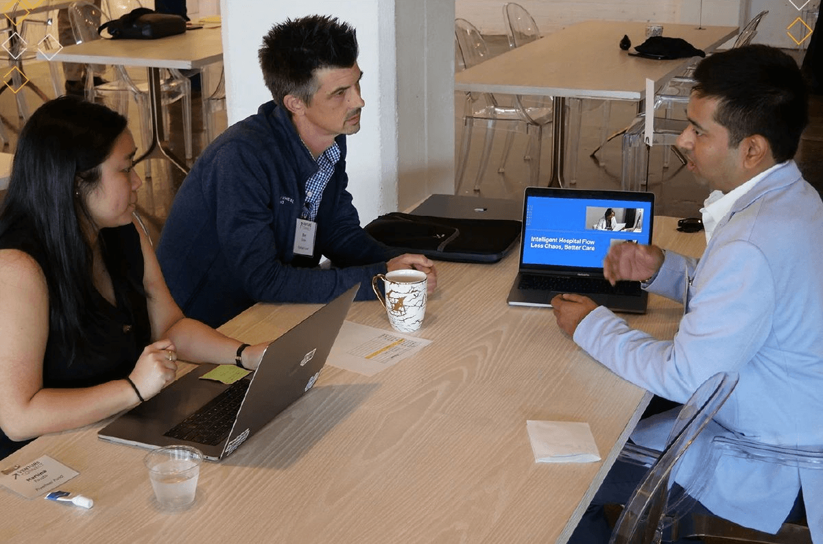 Three people sit at a table in a meeting room, engaging in a discussion with laptops and a tablet.