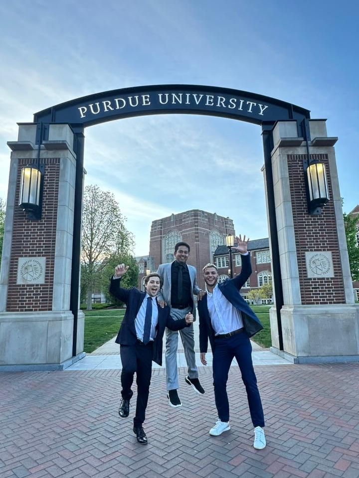 Three people in suits and casual clothing celebrating under Purdue University archway, with buildings and trees in background.