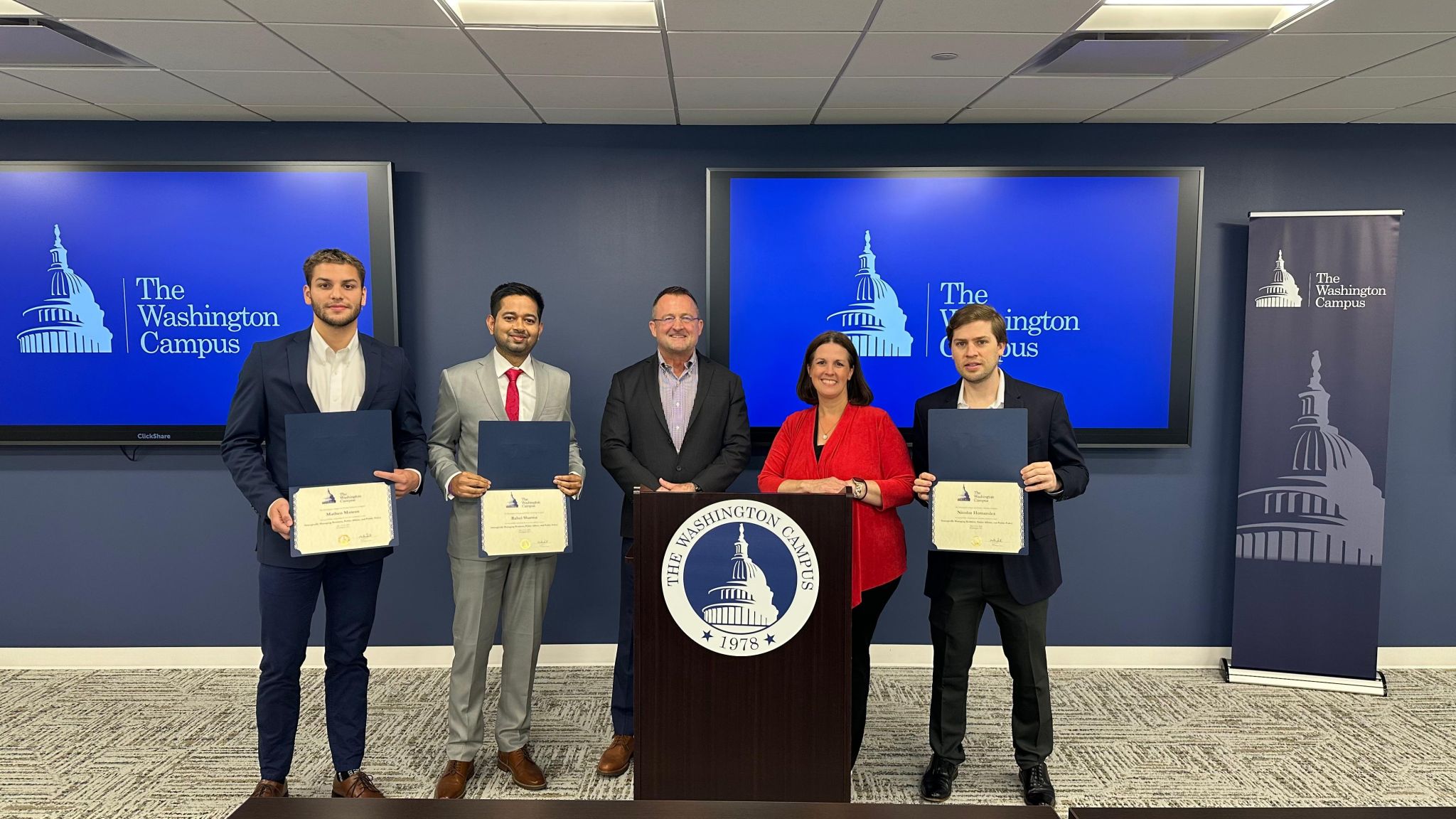 Five people standing behind a podium with a seal, holding certificates, in a conference room with screens displaying 'The Washington Campus'.