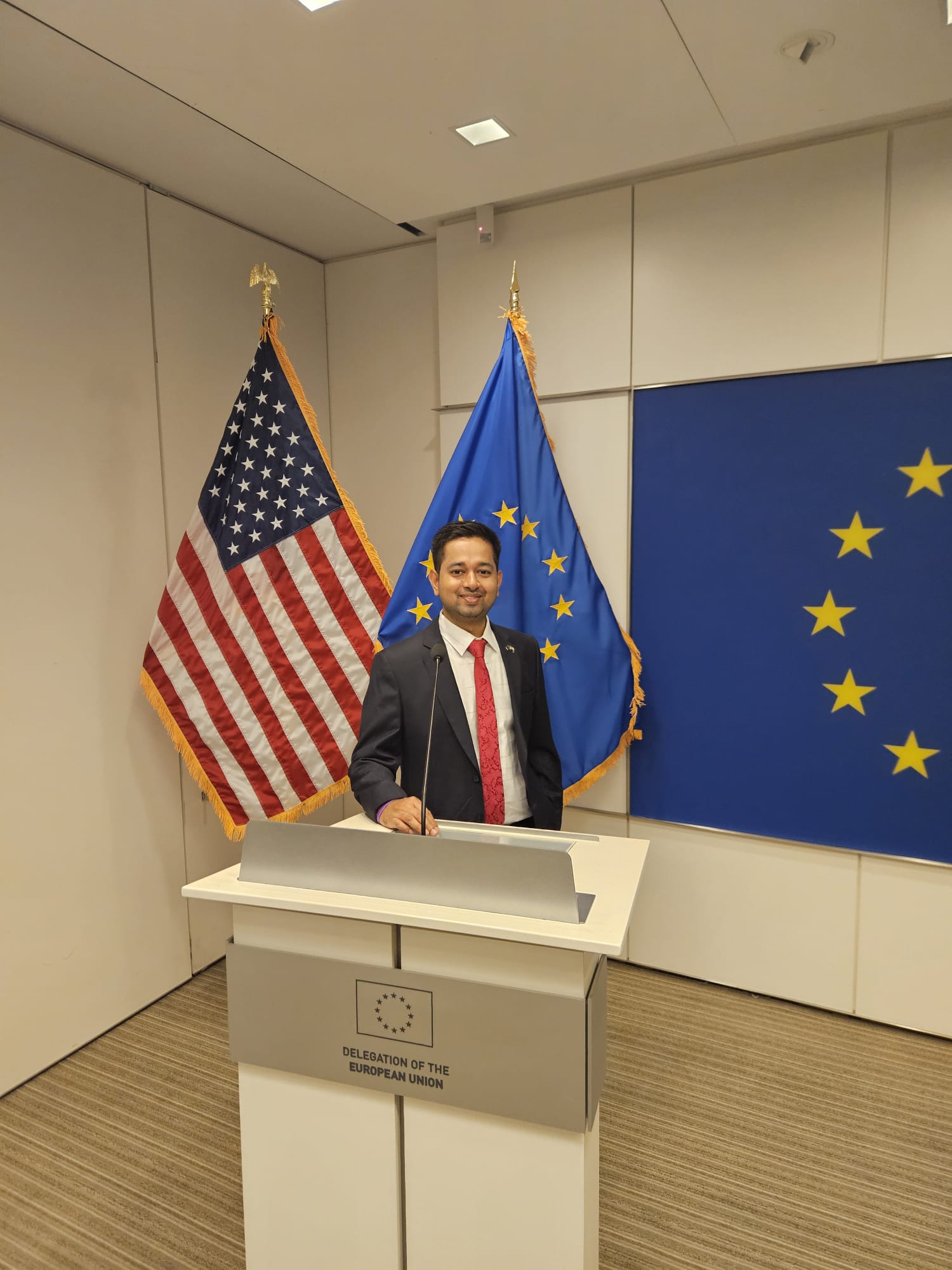 Man standing at a podium with American and European Union flags behind him, in a conference room.