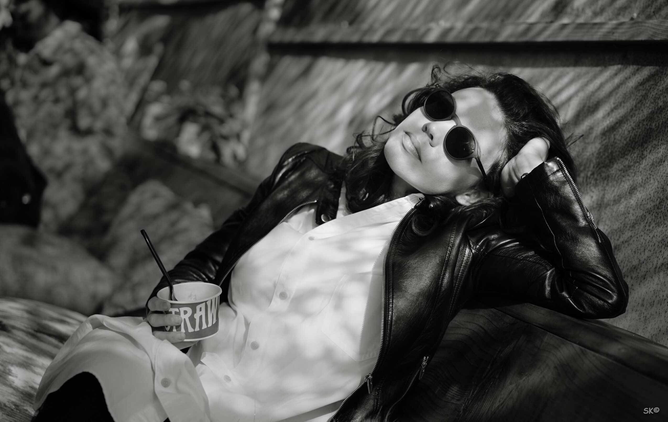 Woman with sunglasses and leather jacket relaxing outdoors, holding a drink with a straw, leaning on a bench.