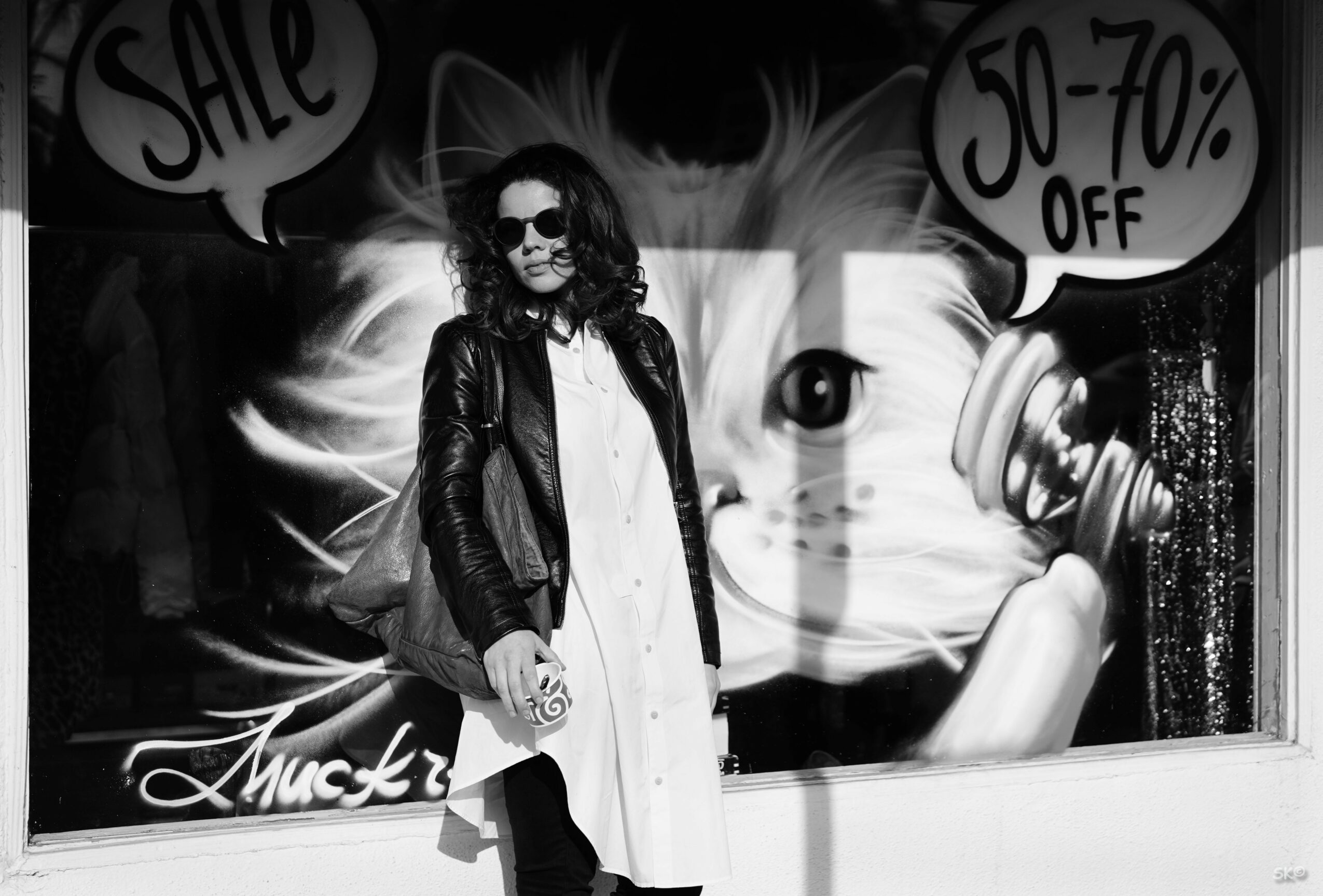 Woman with sunglasses and a leather jacket standing in front of a store window with sale signs and a large cat face image.