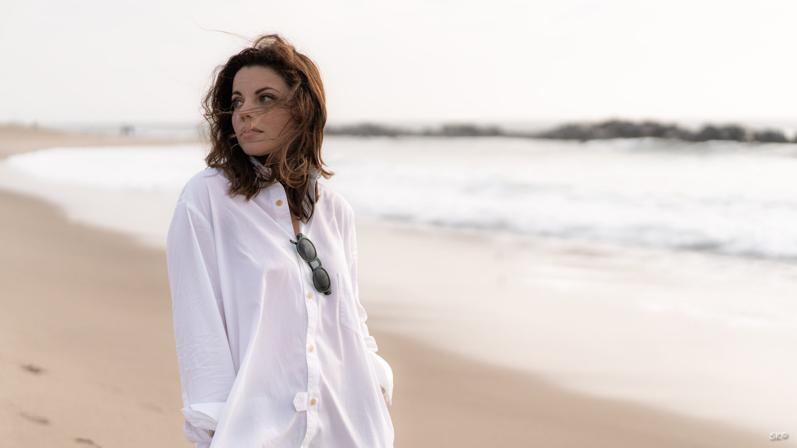 Woman with curly hair wearing a white shirt standing on a beach with ocean waves in the background.