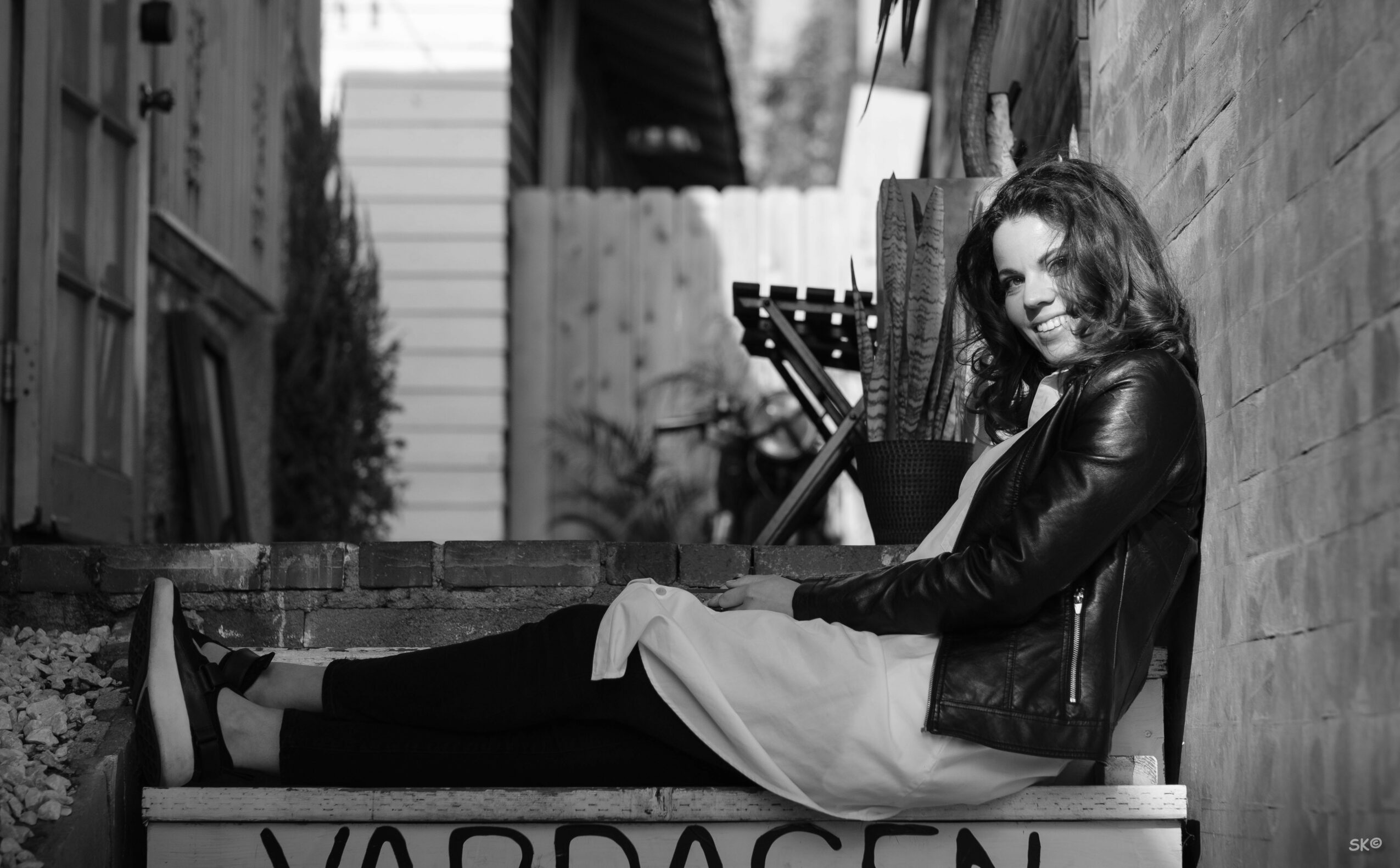 Woman sitting on a ledge, leaning against a brick wall, smiling, wearing a leather jacket and dress, in an outdoor setting.
