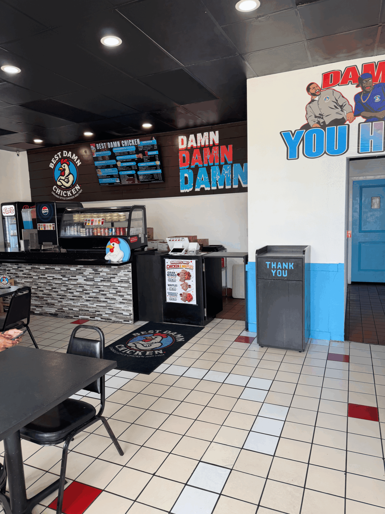 Diner interior with counter, menu, and seating area, featuring colorful signs and a trash bin, tiled floor, and a blue door.