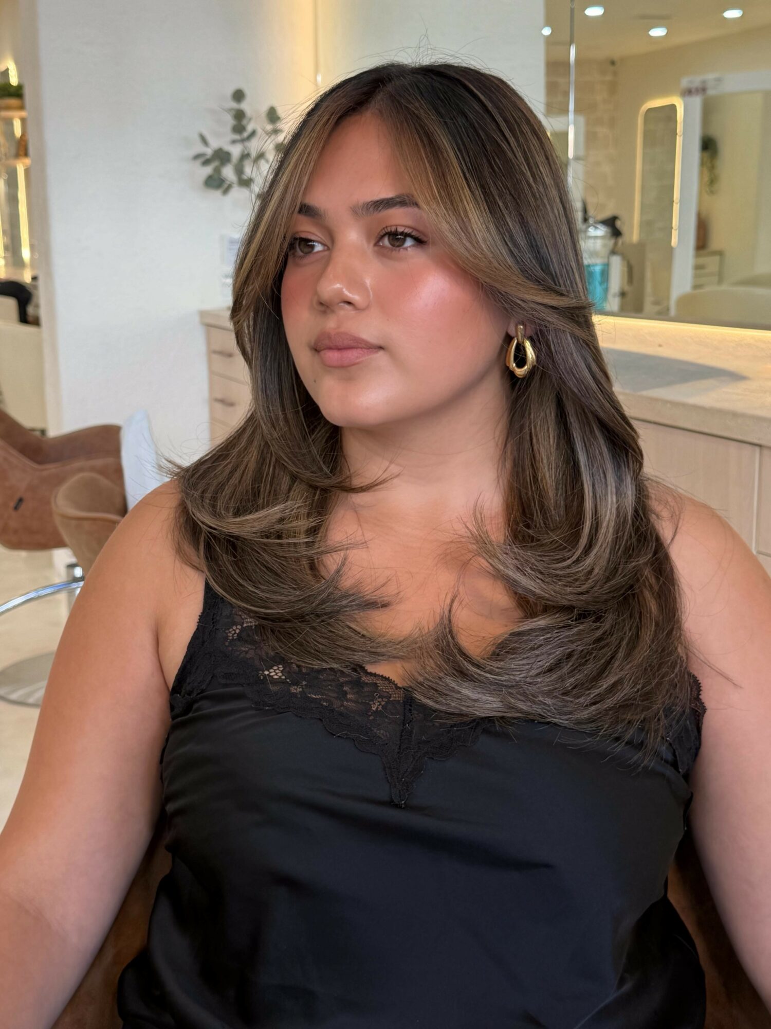 Woman with long, wavy hair and gold hoop earrings, wearing a black top, sitting indoors with a neutral expression.