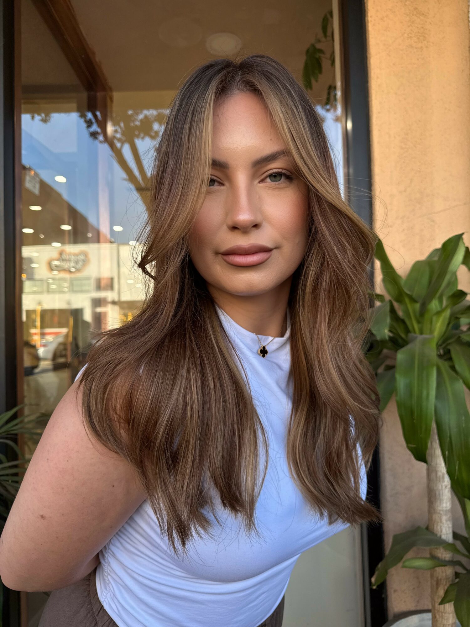 Young woman with long, wavy hair standing outdoors near a plant, wearing a light blue top, smiling softly.