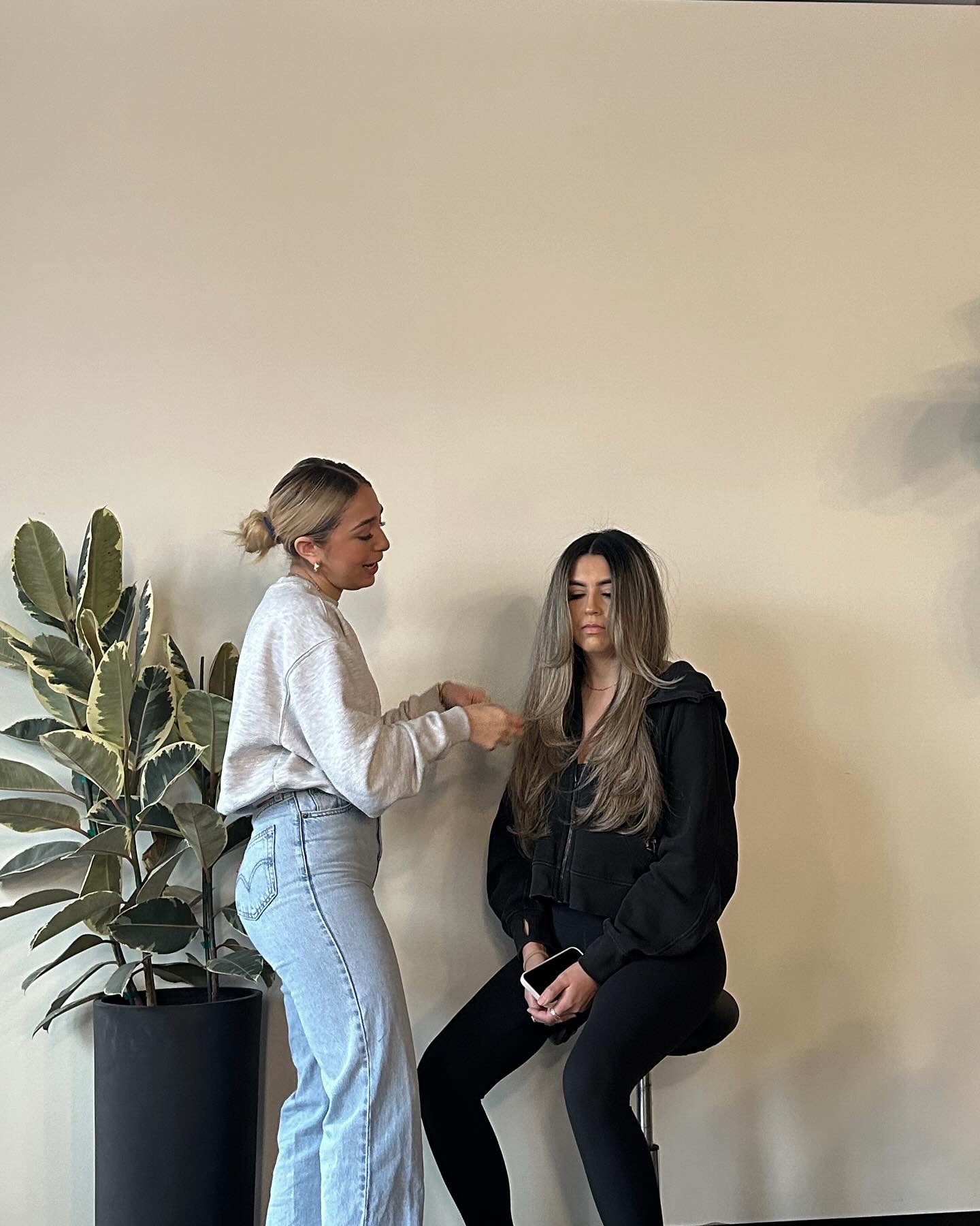 Two women sitting against a plain wall, one with blonde hair in a bun, the other with long hair, wearing dark clothing.
