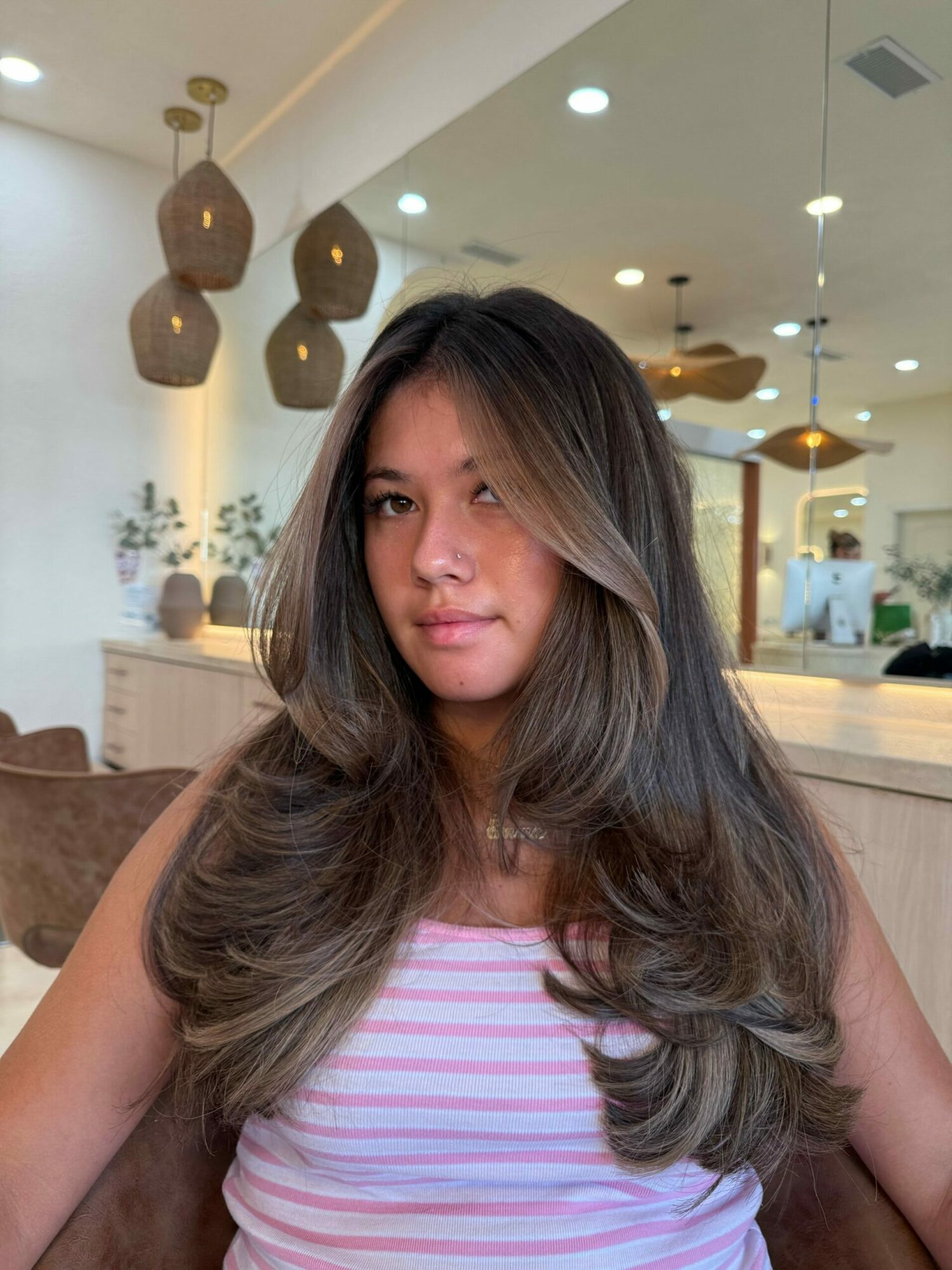 Woman with long wavy hair wearing a pink and white striped shirt, sitting indoors with modern decor in background.