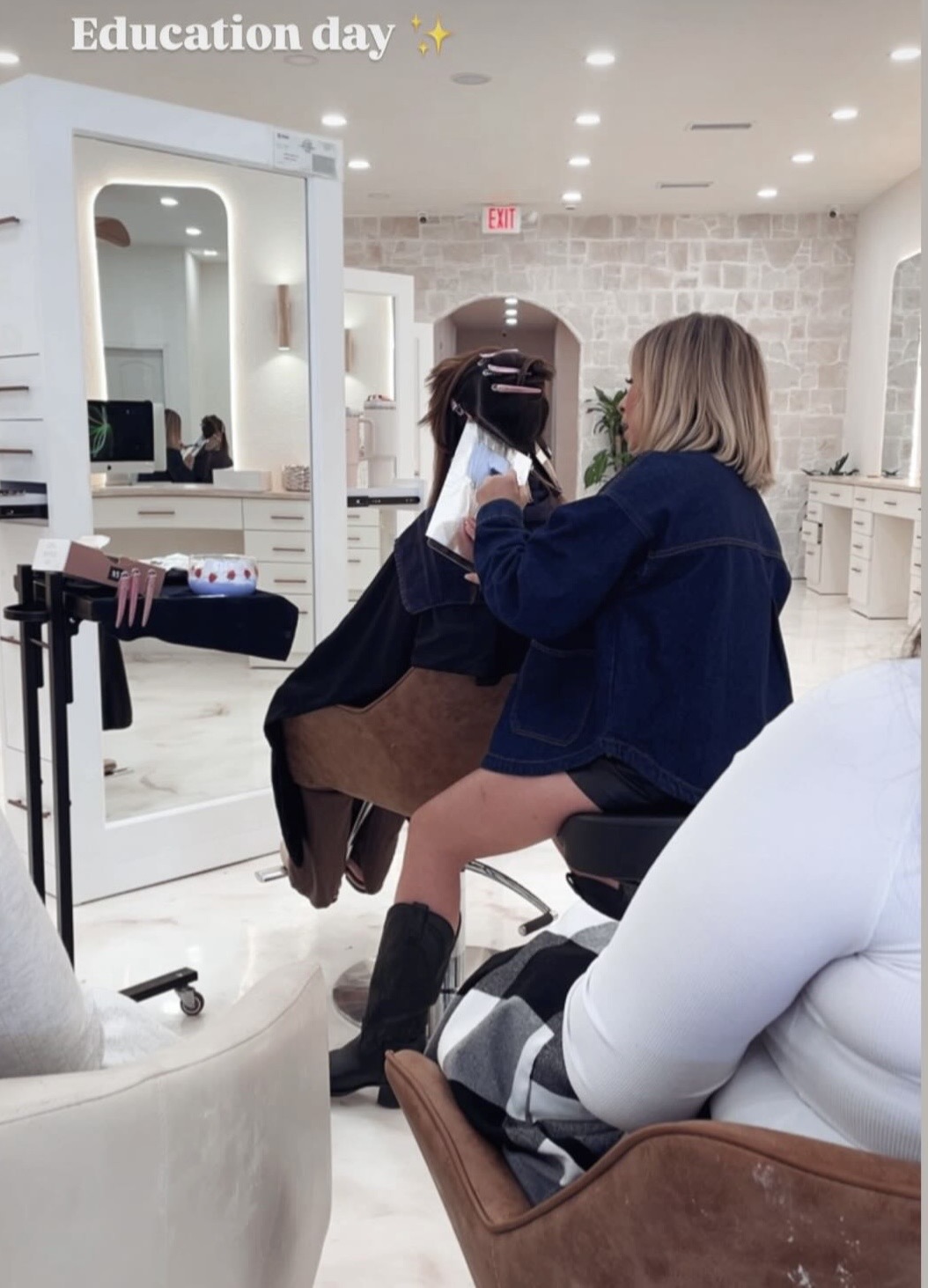 A person receives a haircut in a salon, with another person sitting nearby, in a well-lit room with mirrors and white decor.